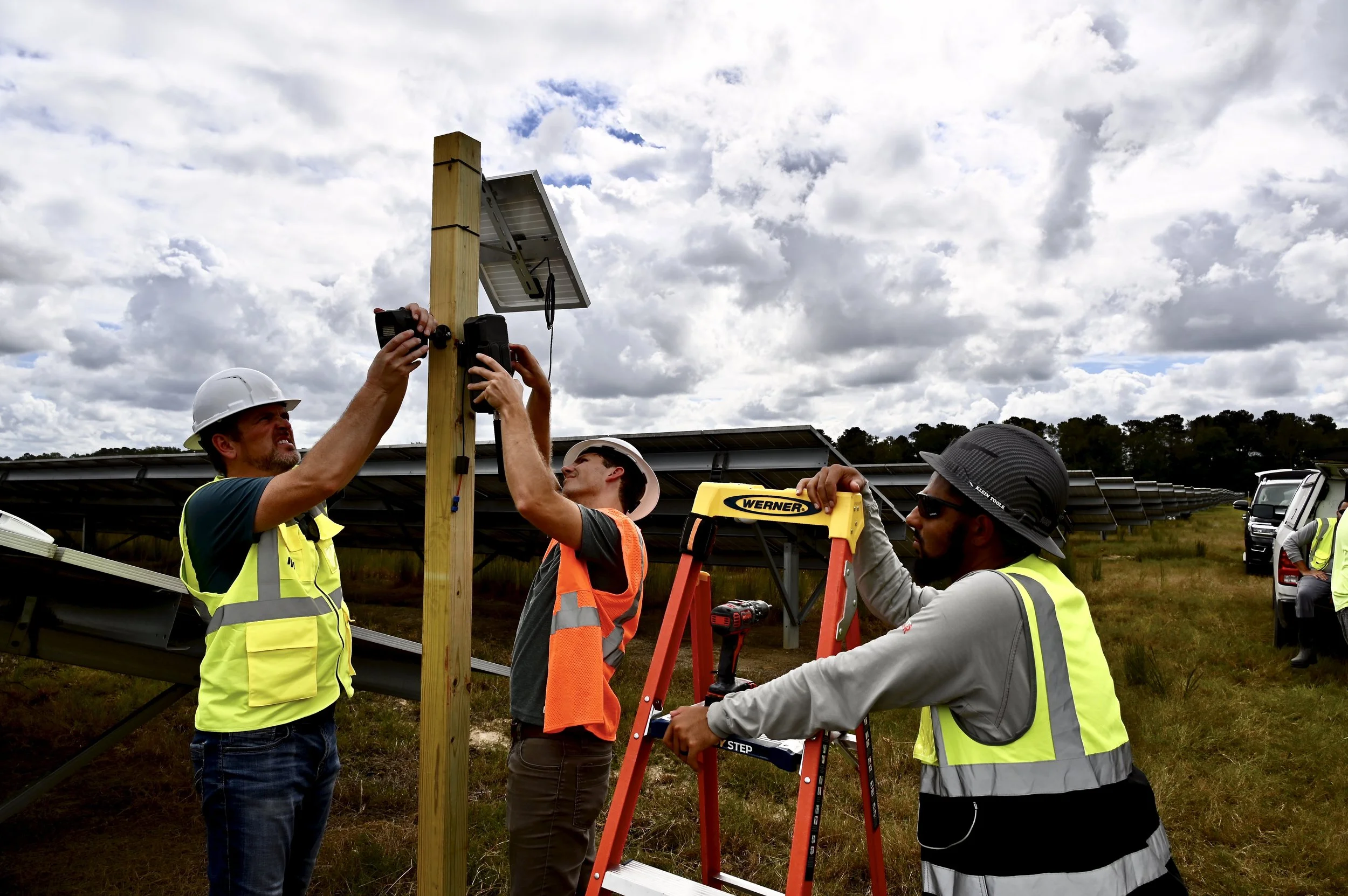 Three workers in safety vests and helmets installing RapidDeployBundle™ camera solution in a solar farm, with a cloudy sky in the background.
