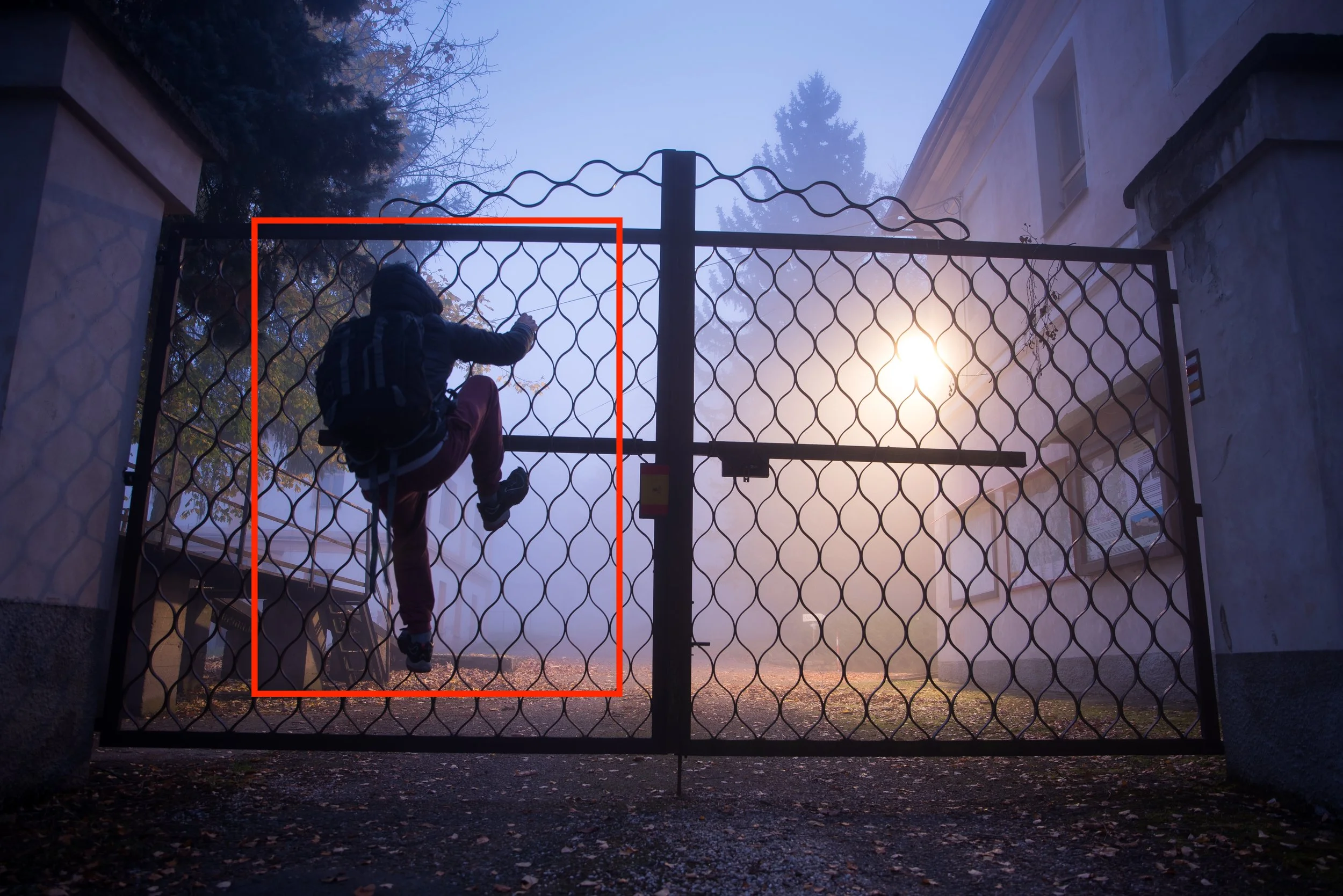 A person wearing a backpack climbing over a closed metal gate early in the morning with fog and sunlight in the background.