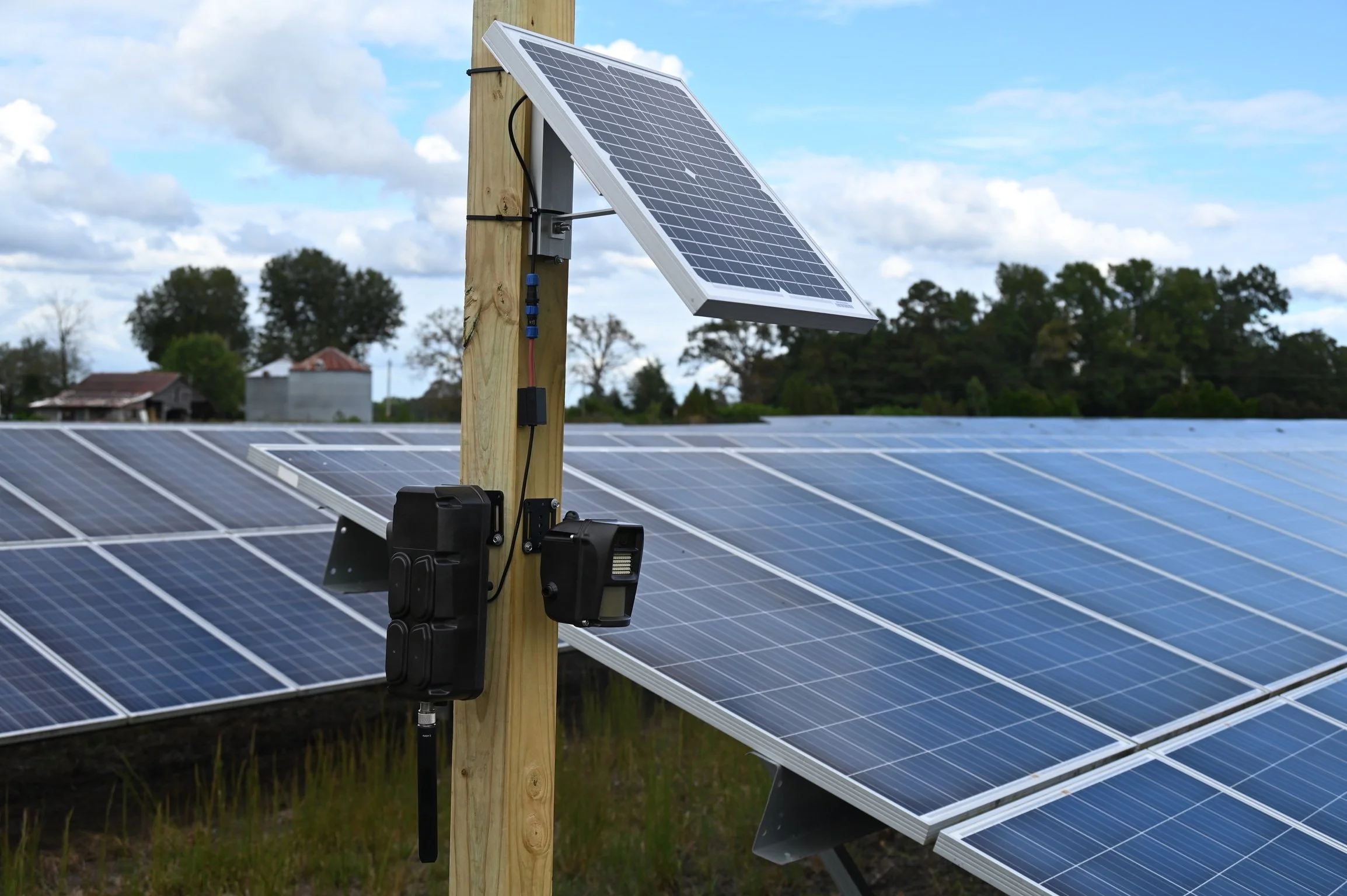 Solar panels with a mounted RapidDeployBundle™ camera solution, in a farmland with trees and buildings in the background.