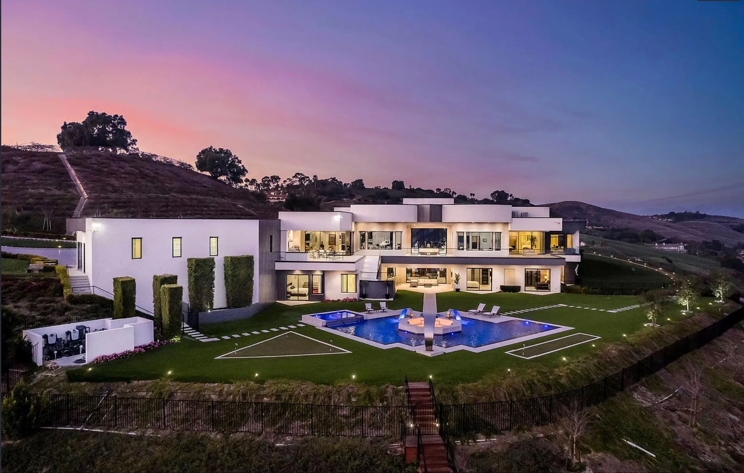 A modern multi-story house with large glass windows and a private swimming pool with lounge chairs, set on a hillside at dusk with the sky showing a purple and pink hue.