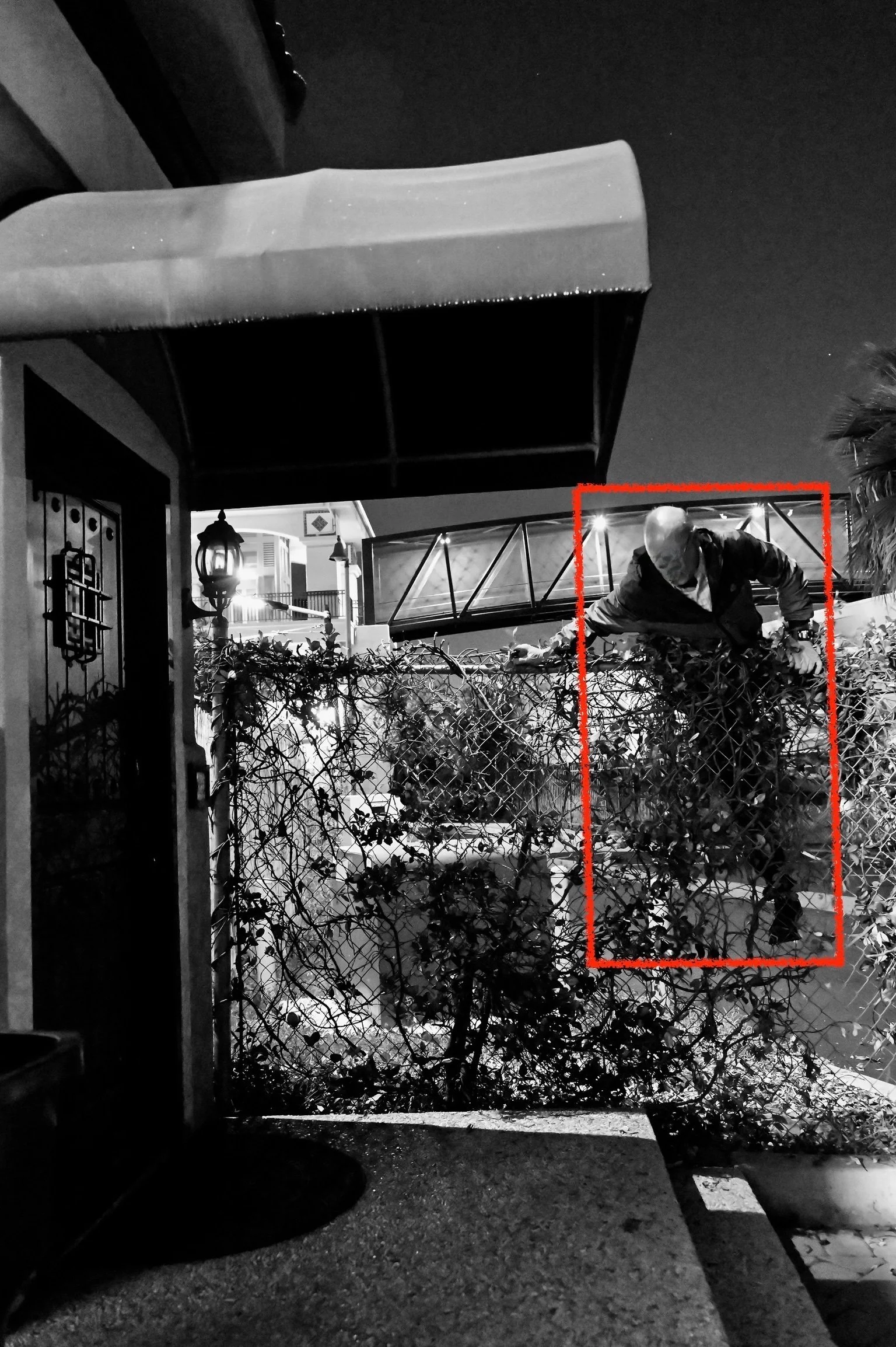 Man in dark clothing climbing over a chain-link fence at night, with houses and outdoor lighting visible in the background.