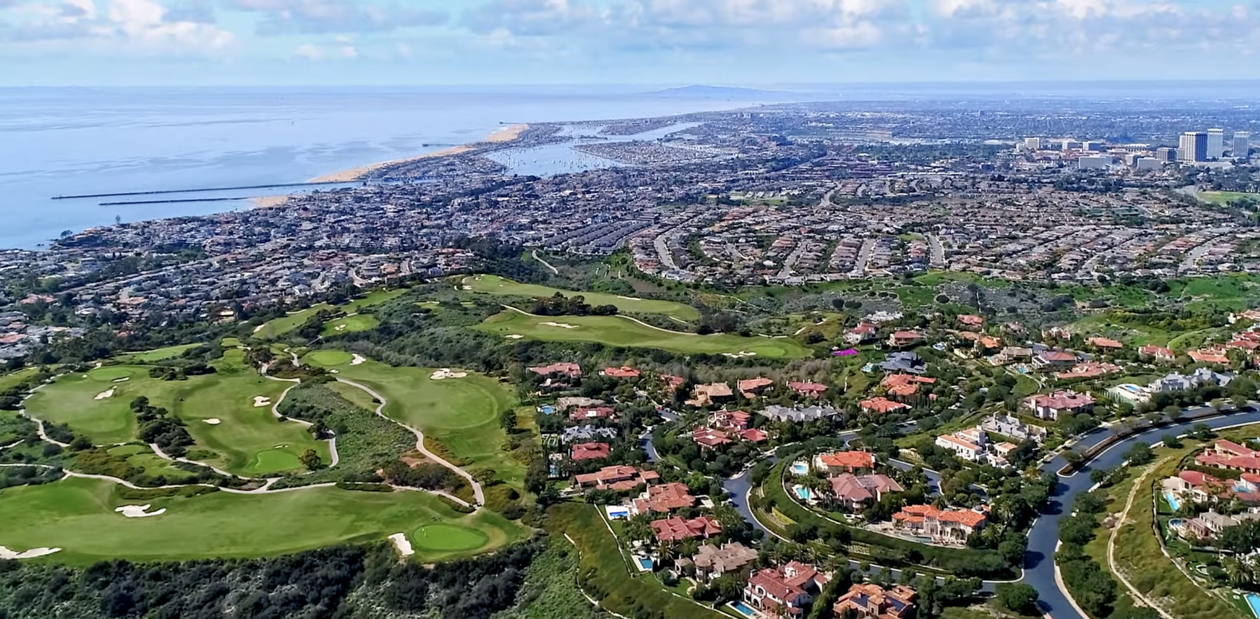 Aerial view of a coastal city with a golf course, residential neighborhood, and shoreline with ocean and harbor in the distance.