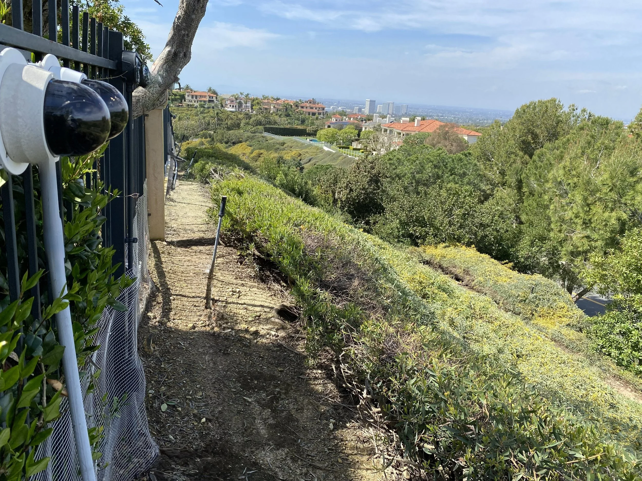 Long‑Range Cameras mounted on a fence over a coastal hillside with a dirt trail, green bushes, trees and a view of distant buildings under a partly cloudy sky.