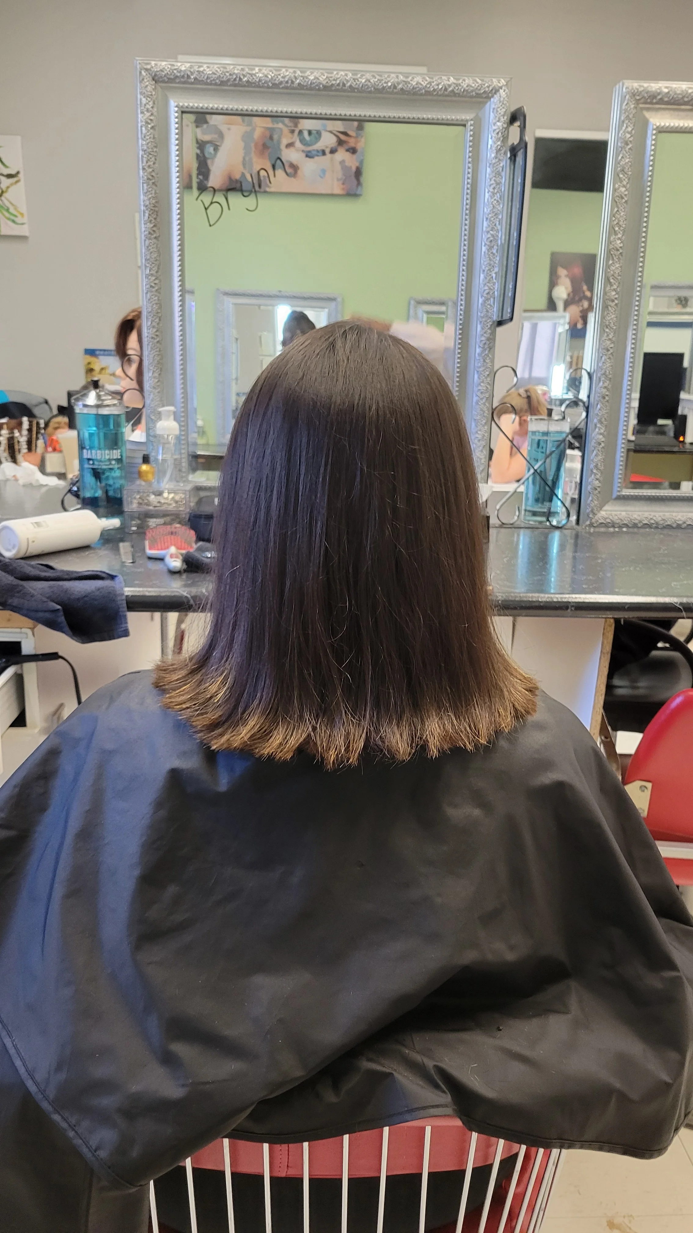 The back of a woman's head with shoulder-length dark hair, sitting in a salon chair with a black cape. In the background, a salon mirror reflects another woman with red hair, and various hair tools are on the counter.