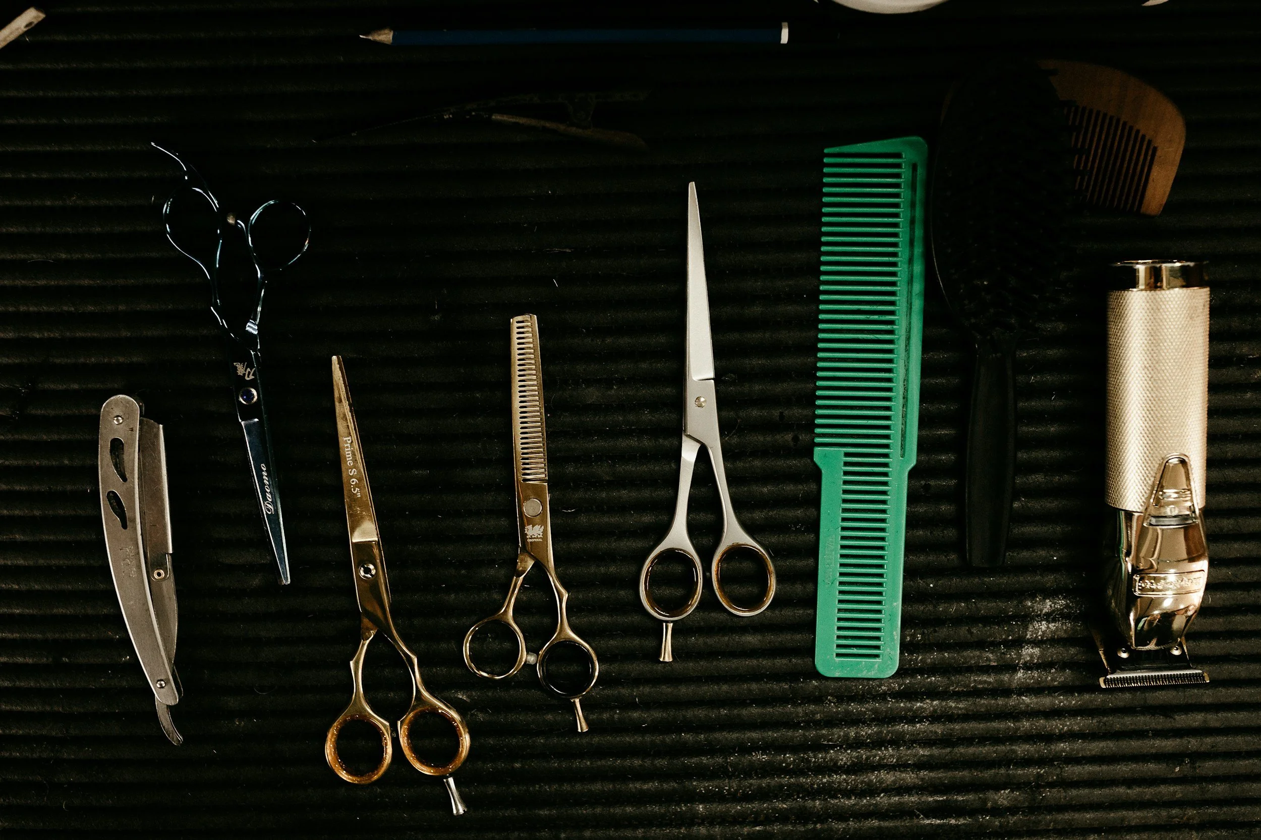 A collection of barber tools including scissors, clipper, razors, combs, and a hairbrush arranged on a black ridged surface.