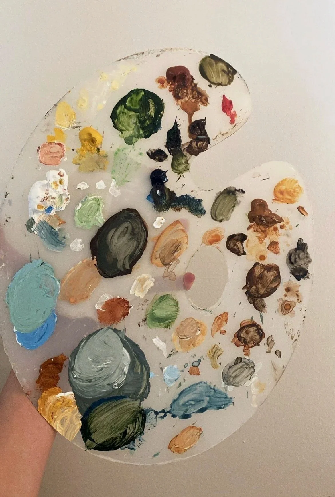 A painter's palette with various mixed and unmixed oil paint blobs in different colors, held against a plain wall.