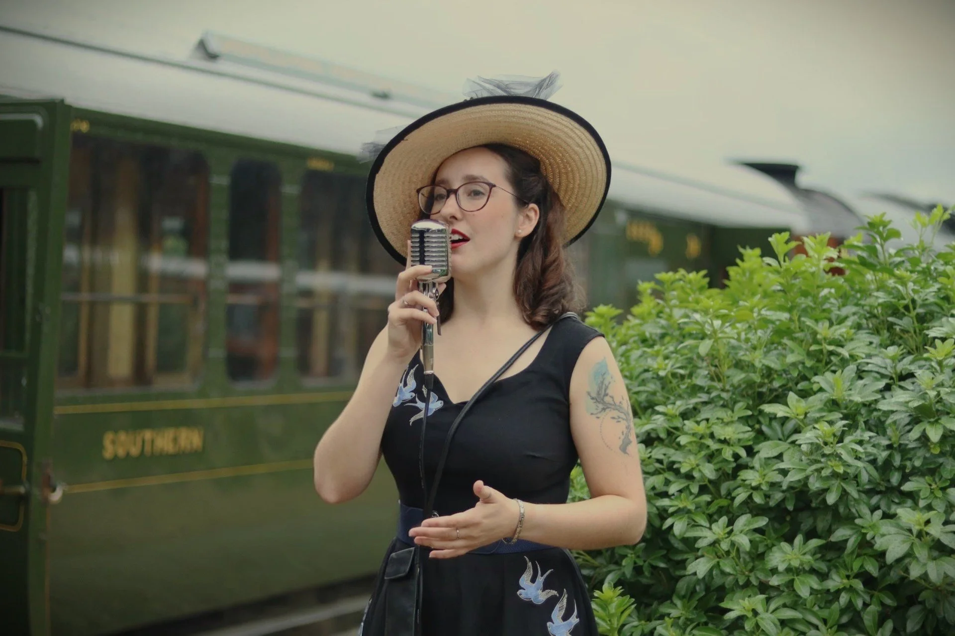 A woman wearing a large sunhat and glasses singing into a vintage microphone outdoors, with a green train and lush green bushes in the background.
