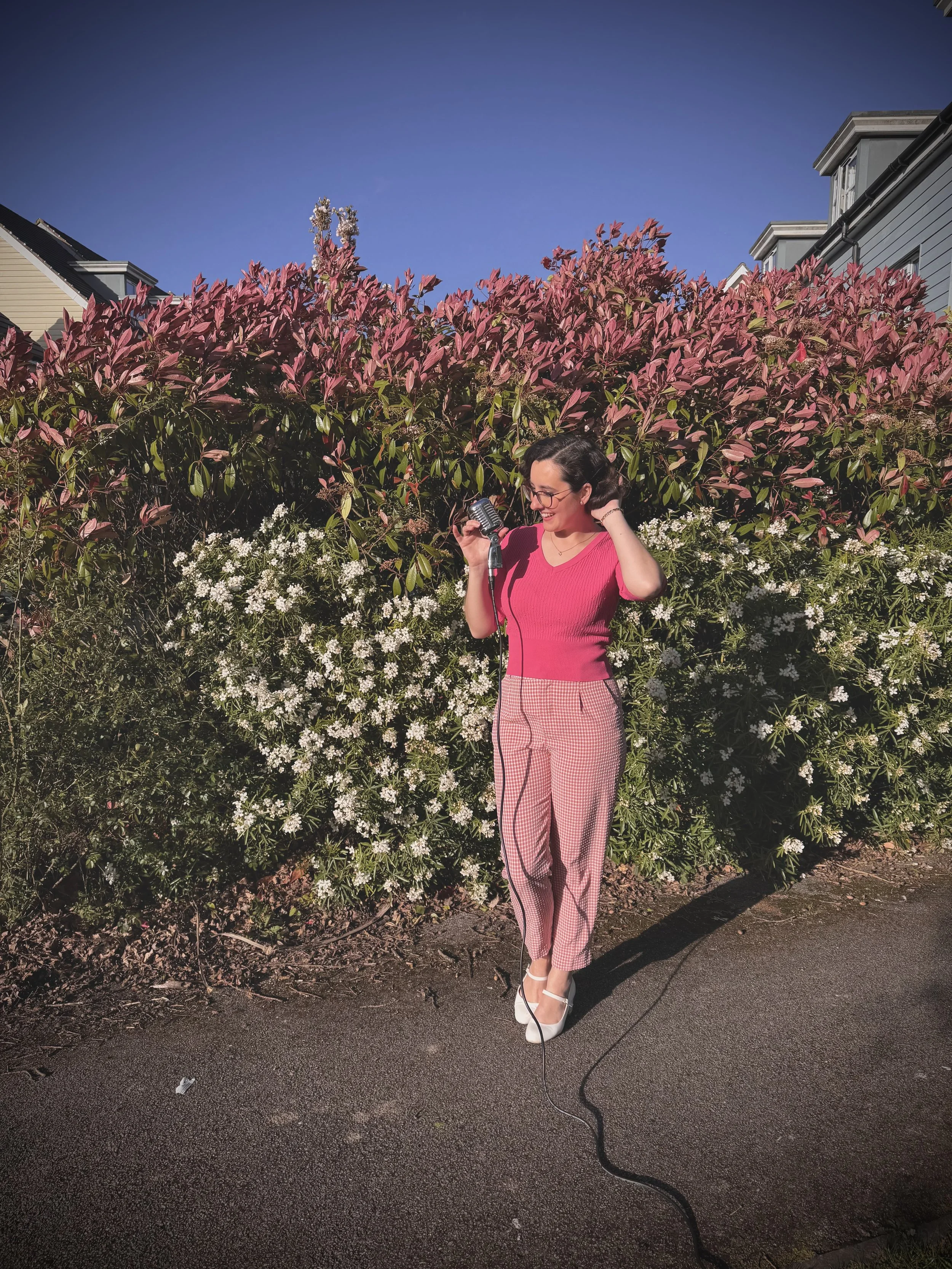 Keziah vintage jazz vocalist in a bright pink top and pink plaid trousers stands on a pavement holding a vintage microphone, smiling, with pink flowering bushes and residential buildings behind her under a clear blue sky. Photo was taken during Sprin