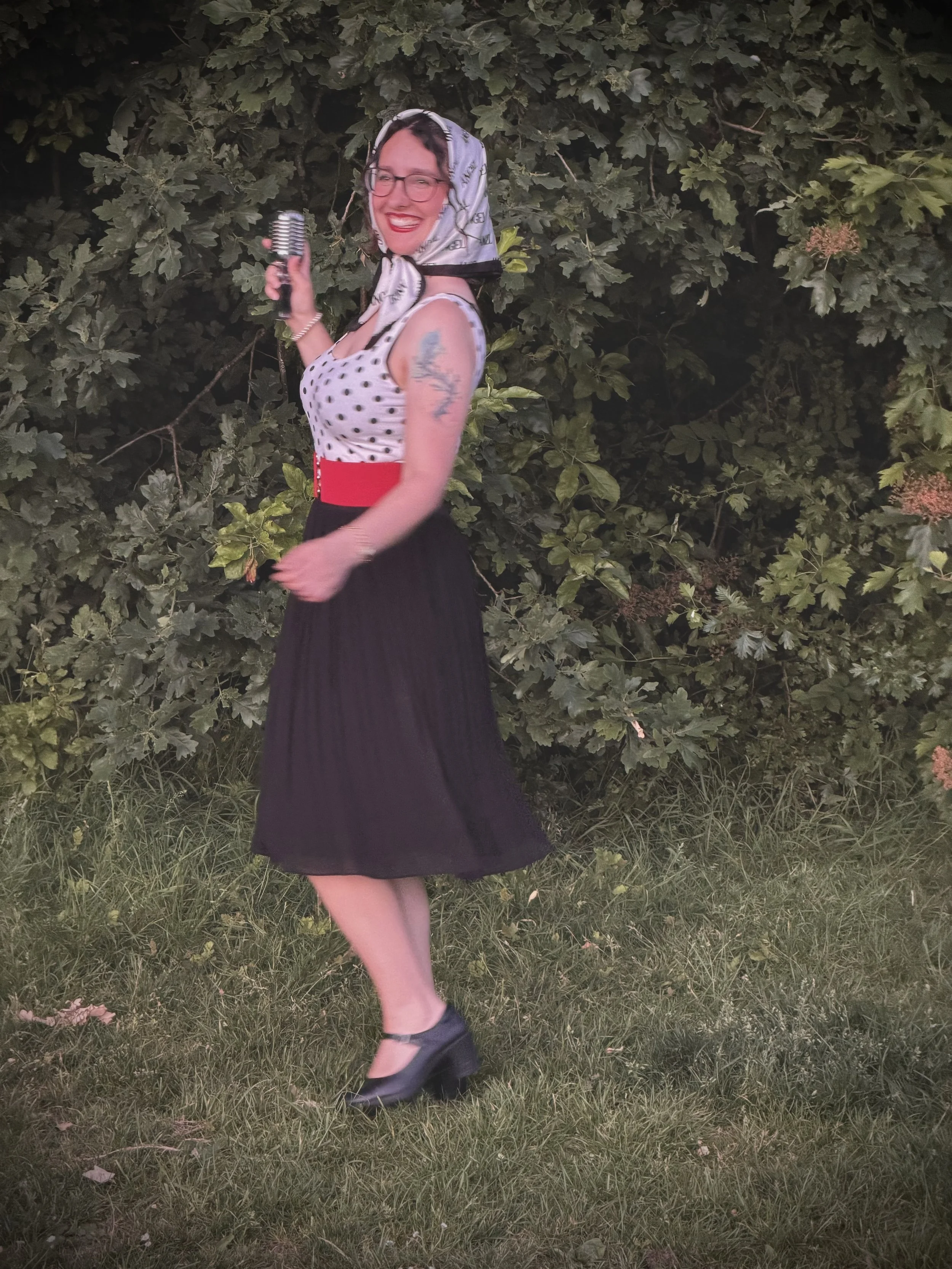 Keziah vintage jazz vocalist smiling and holding a vintage microphone, standing in front of a leafy green bush at night. She is wearing glasses, a polka dot top, a black skirt with a red belt, and black shoes, with a headscarf on her head and a tatto
