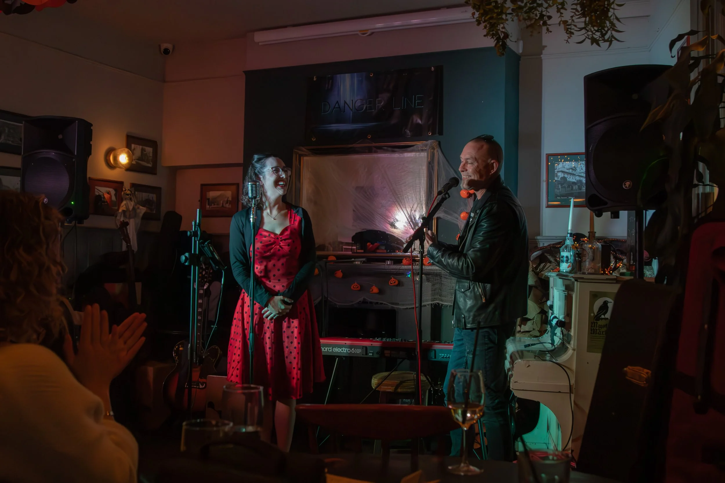 A man and woman performing on stage at a cozy venue decorated with Halloween-themed ornaments, including small pumpkins. The woman is wearing a red polka dot dress and glasses, smiling, while the man is dressed in a black leather jacket, singing into