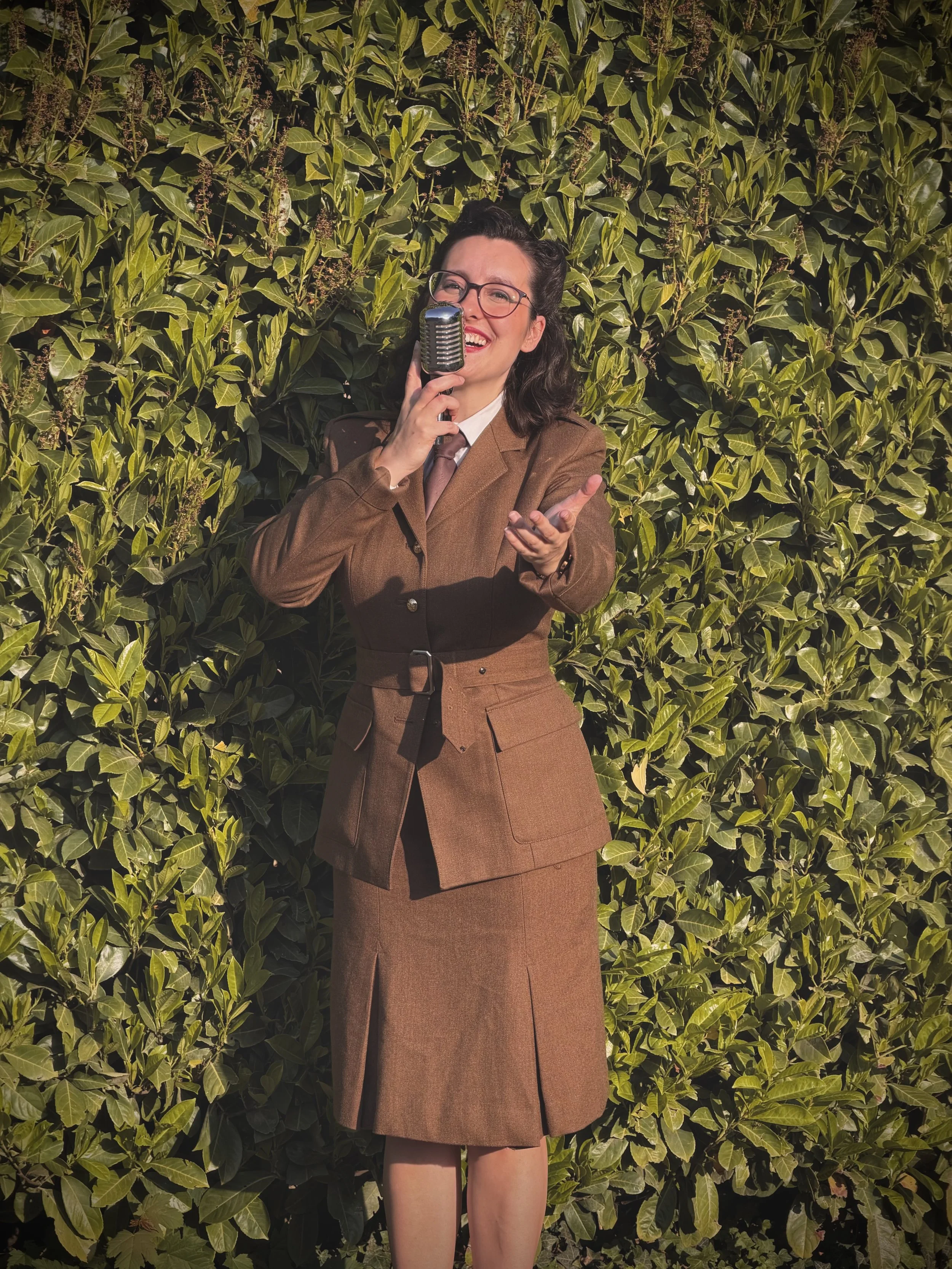 Keziah vintage jazz vocalist with dark hair and glasses singing into a vintage microphone outdoors in front of a lush green hedge. She is wearing the ENSA (Entertainment Services Association) uniform that Dame Vera Lynn wore during World War 2.