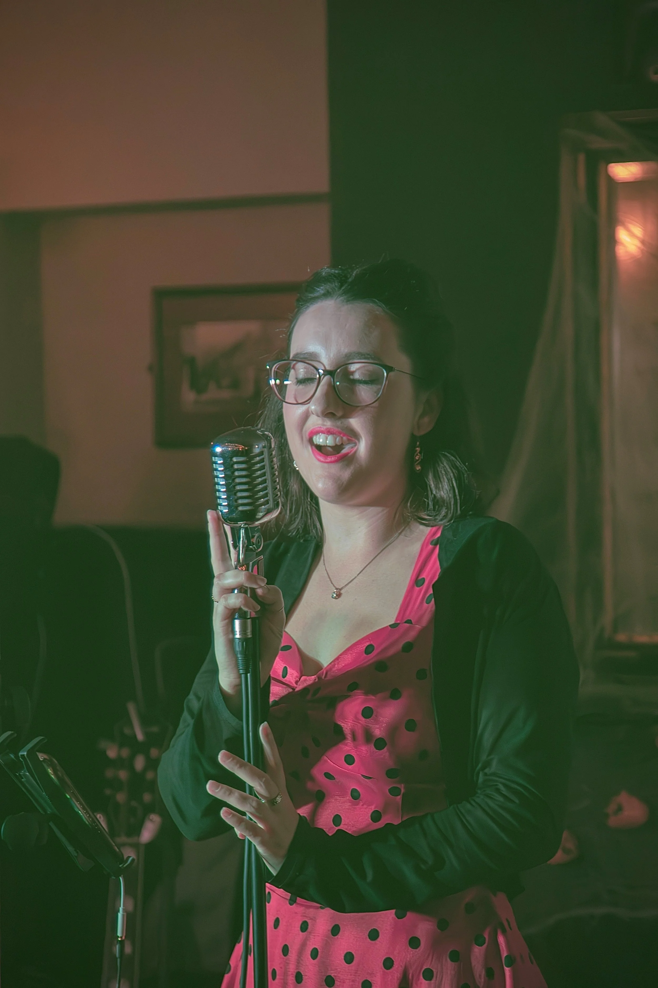 A woman with glasses and dark hair singing or speaking into a vintage microphone, wearing a pink polka dot dress with a black jacket in a dimly lit room.