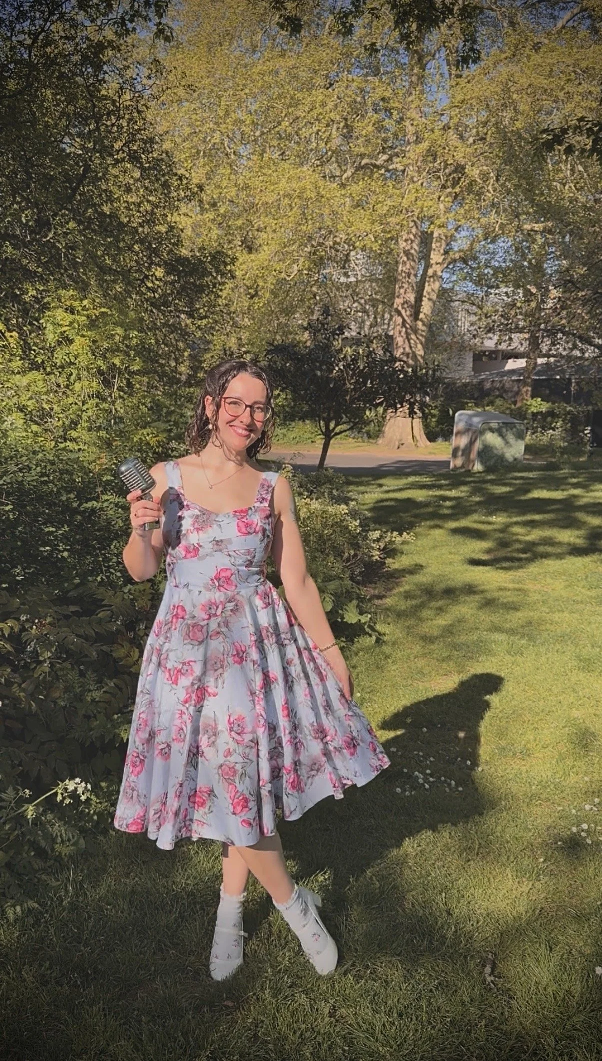 Keziah vintage jazz vocalist wearing a floral dress and white heels holding a vintage microphone, standing outdoors on a sunny day with green grass and trees in the background.