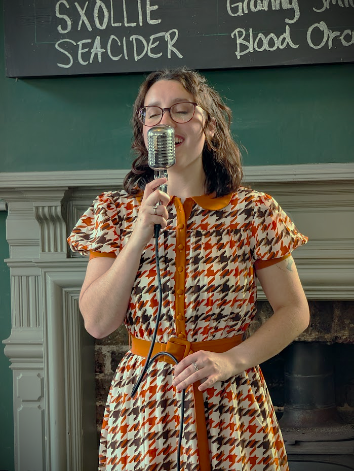 A woman with glasses and wavy brown hair, wearing a multicolored houndstooth dress, is standing in front of a fireplace and holding a vintage microphone, smiling with her eyes closed. A chalkboard behind her displays menu items such as 'SXOLLIE SEACI