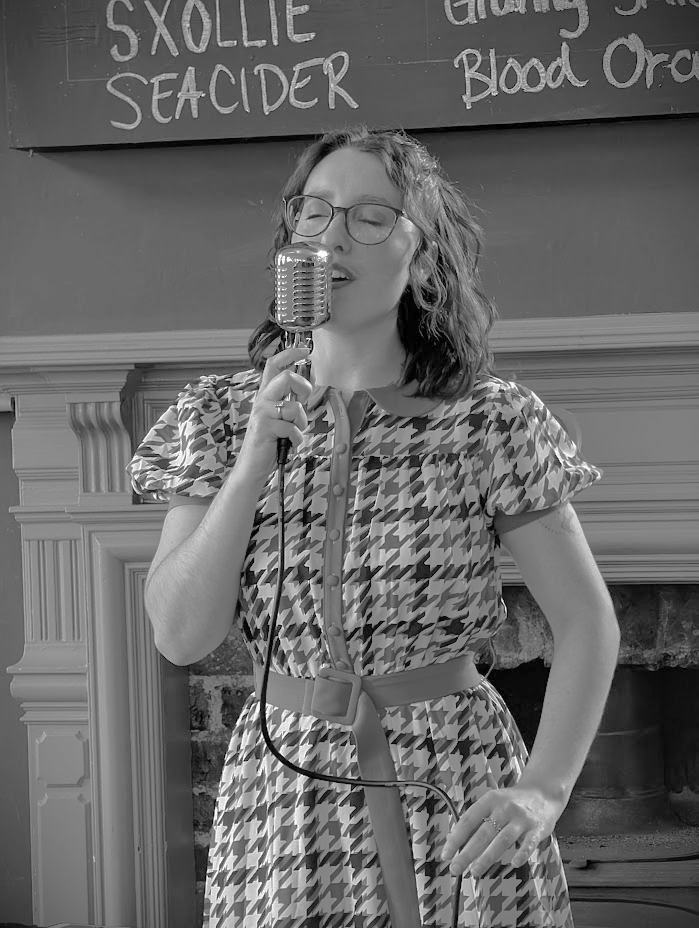 A woman singing into a vintage microphone in a live performance setting. She is wearing glasses and a patterned dress, standing in front of a fireplace with a chalkboard menu above.