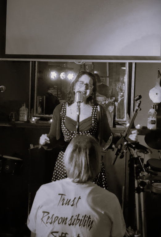 A woman with glasses singing into a microphone with a person sitting in front, in a dimly lit room with musical equipment and a window in the background