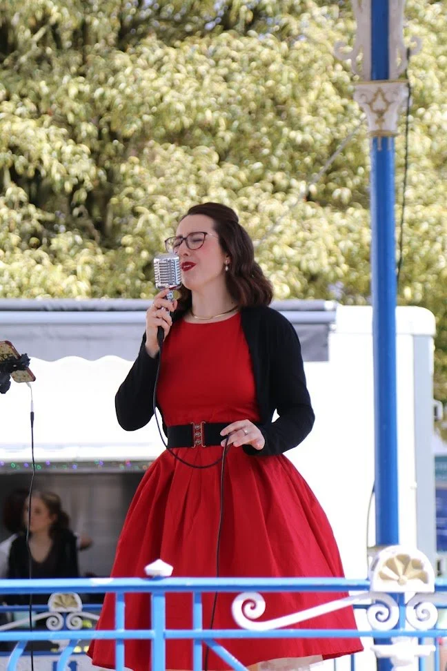 Woman singing into a vintage microphone outdoors, wearing a red dress and black cardigan, with green trees in the background.