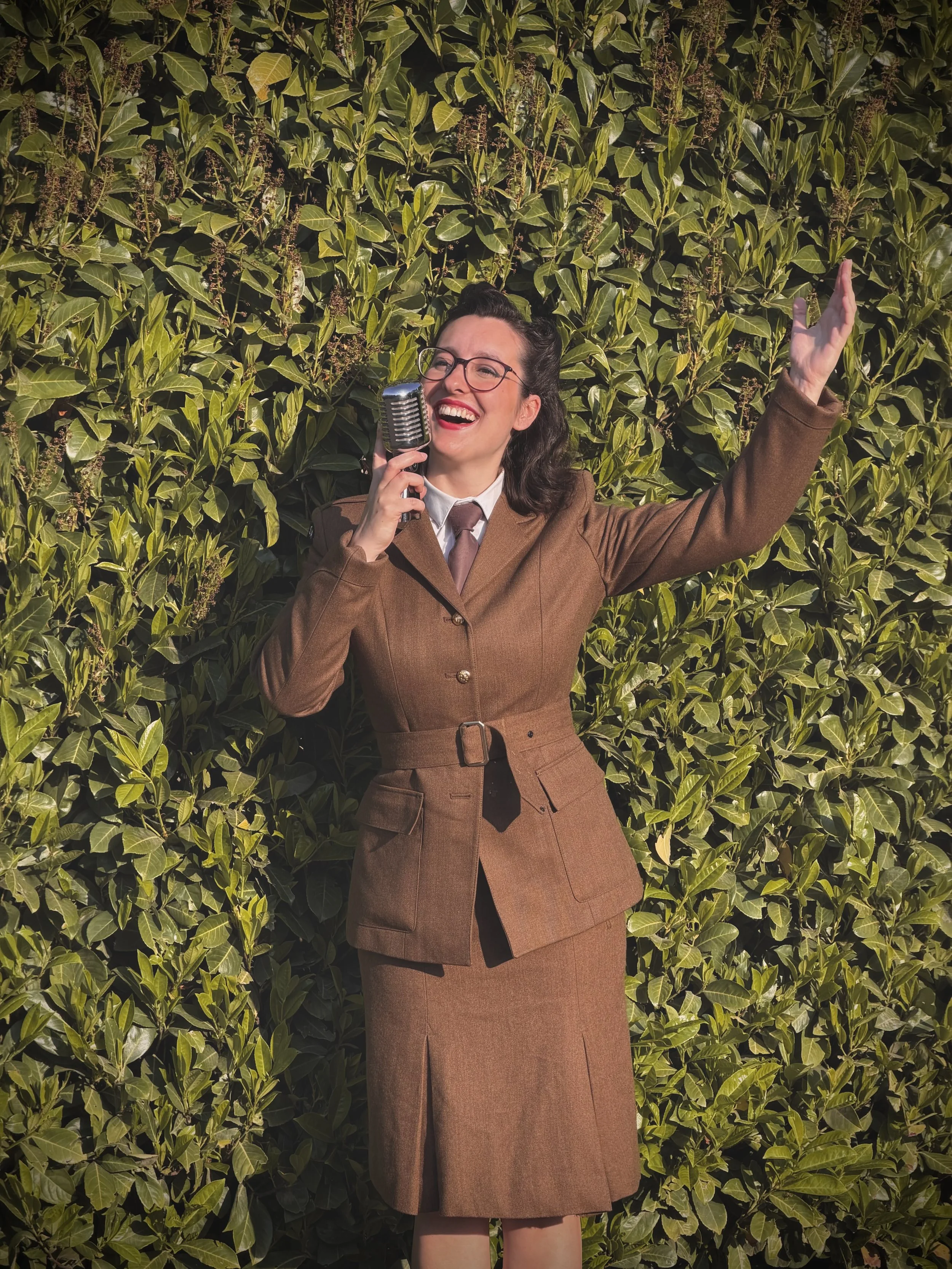 Keziah vintage jazz vocalist with dark wavy hair, glasses, and red lipstick, dressed in a brown vintage suit with a skirt and tie, singing into a retro microphone while standing in front of green leafy bushes. She is wearing an ENSA (Entertainment Se