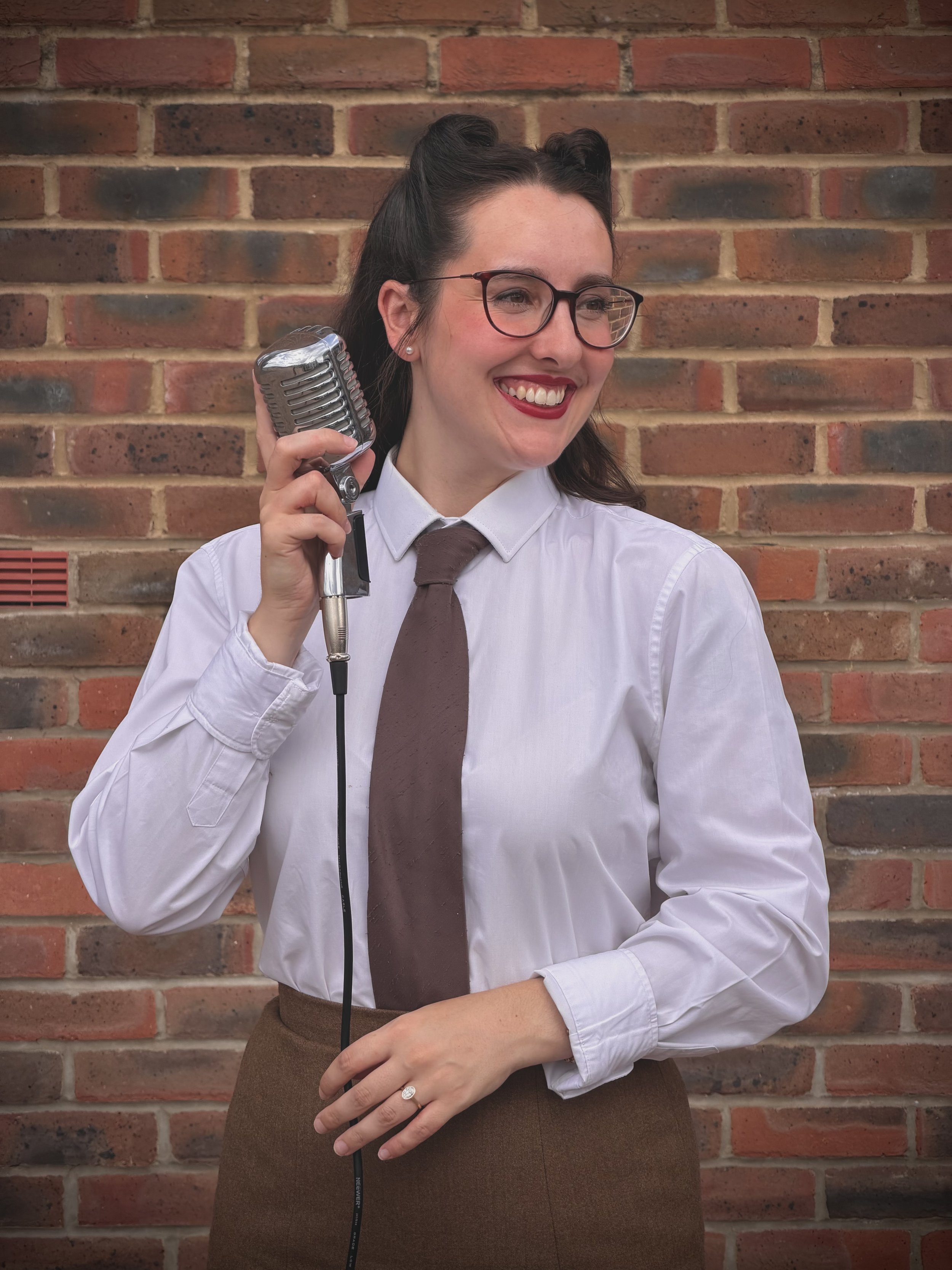 Keziah jazz vocalist is wearing a brown tie and holding a vintage microphone whilst smiling. She is wearing ENSA (Entertainment Services Association) uniform that Dame Vera Lynn wore during World War 2.