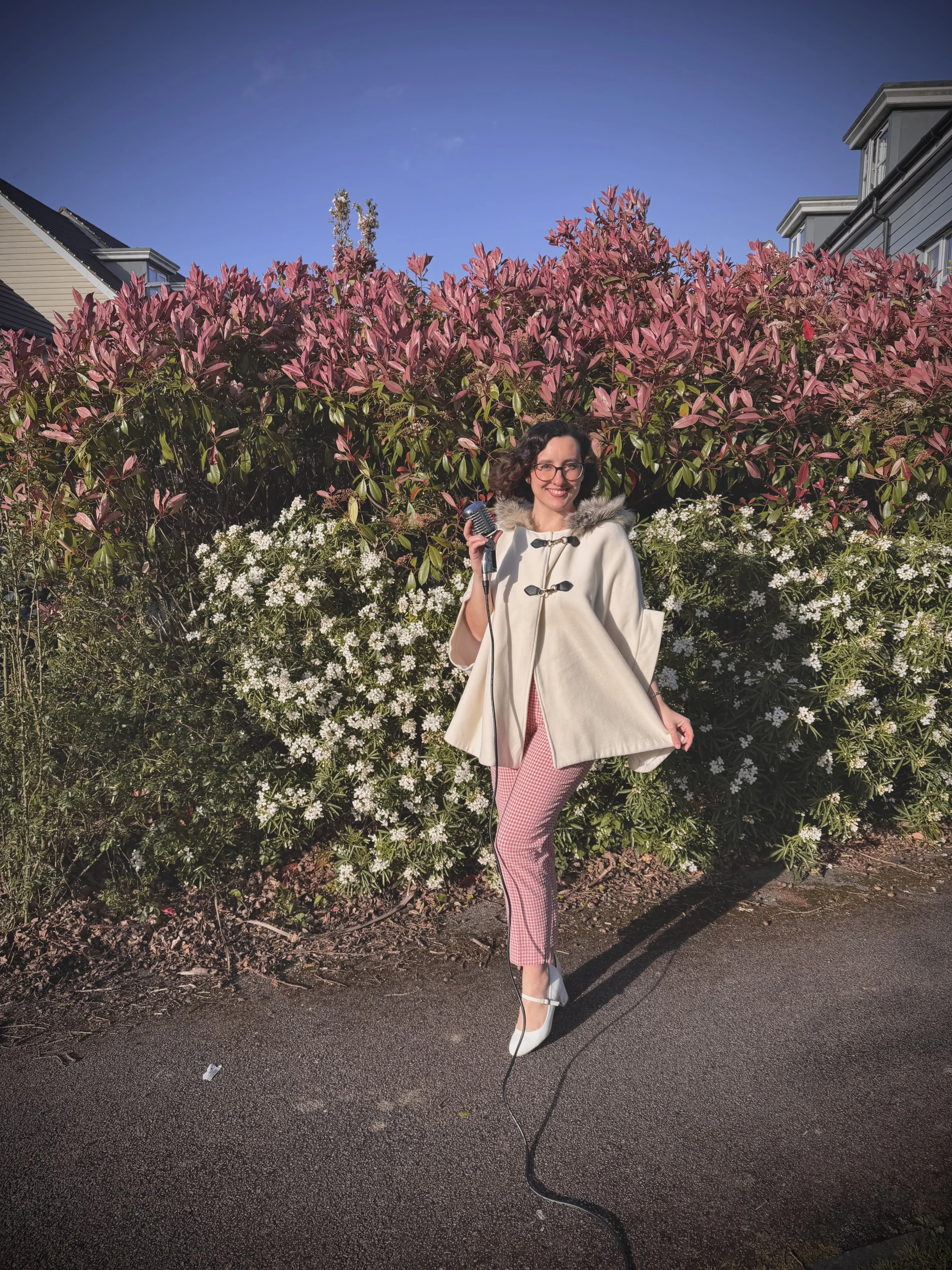 Keziah vintage jazz vocalist with curly dark hair and glasses smiling, holding a vintage microphone, wearing a white coat with fur collar, pink and white checkered trousers, white high heels, standing on a pavement in front of pink flowering bushes a