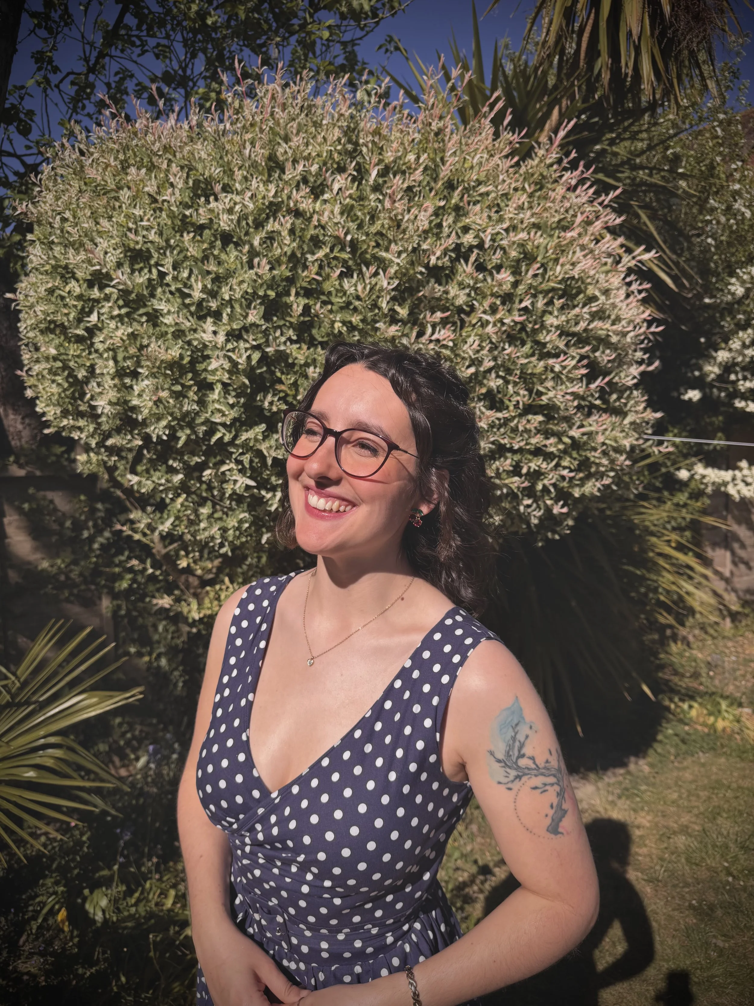 Keziah vintage jazz vocalist with glasses and a polka dot dress smiling outdoors with a large flowering shrub behind her and a clear blue sky.