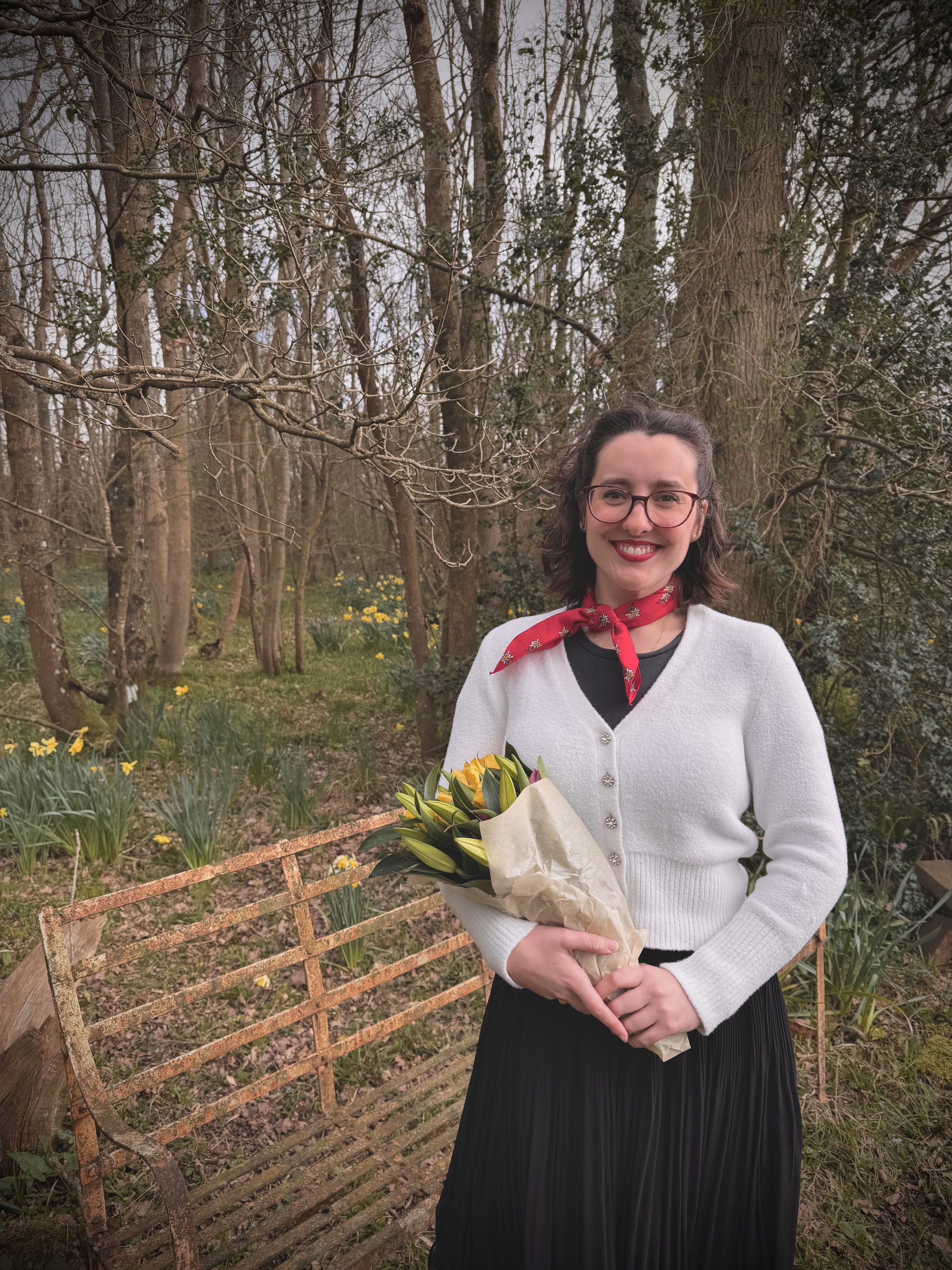 A woman smiling outdoors holding a bouquet of yellow and pink flowers, standing in front of a garden with trees and yellow flowers.