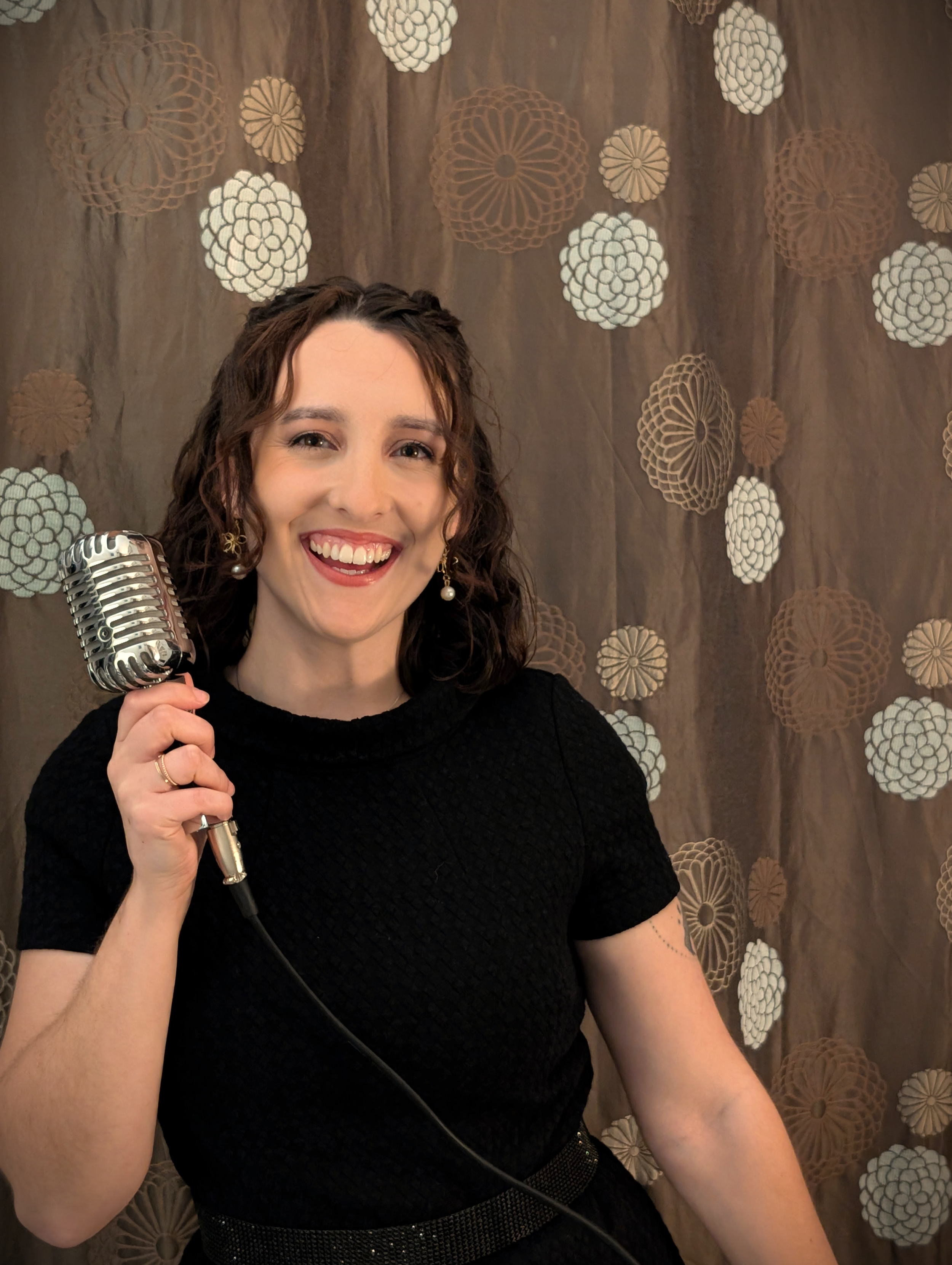 A smiling woman with wavy brown hair holding a vintage-style microphone, wearing a black dress with gold earrings and a black belt, standing in front of a brown patterned curtain with white and brown circular designs.