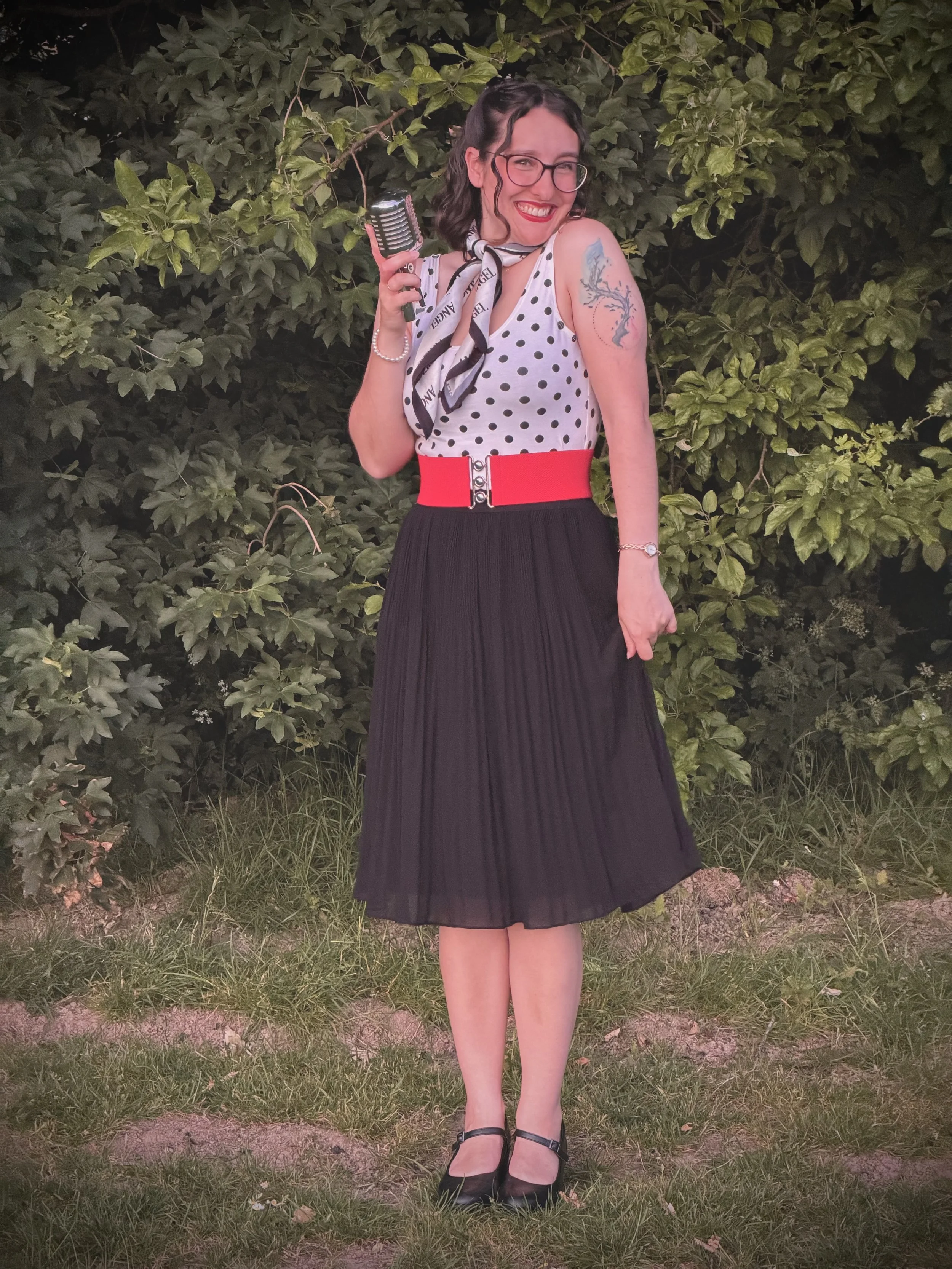 Keziah vintage jazz vocalist wearing a polka-dot top, black skirt, and black heels, holding a vintage microphone, standing outdoors at night in front of leafy bushes, smiling.