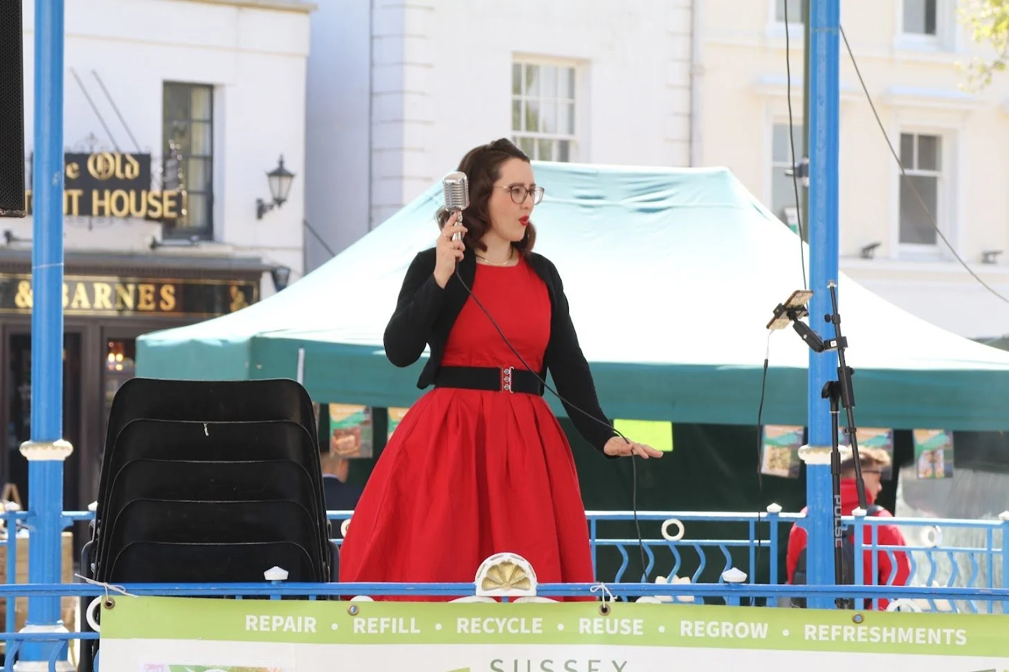 A woman dressed in vintage style red dress and black jacket singing or speaking into a microphone on an outdoor stage, with a tripod holding a phone nearby.