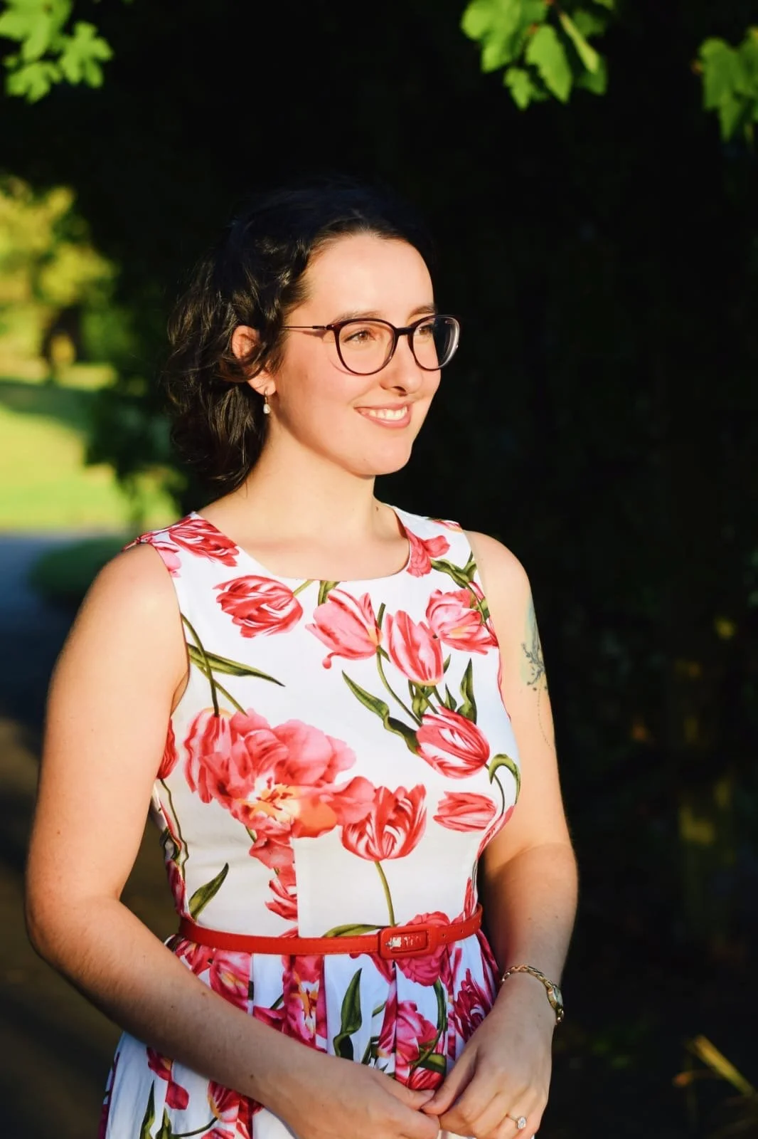 Keziah vintage jazz vocalist in a white dress with pink and red floral pattern, wearing glasses, smiling, outdoors in daylight.