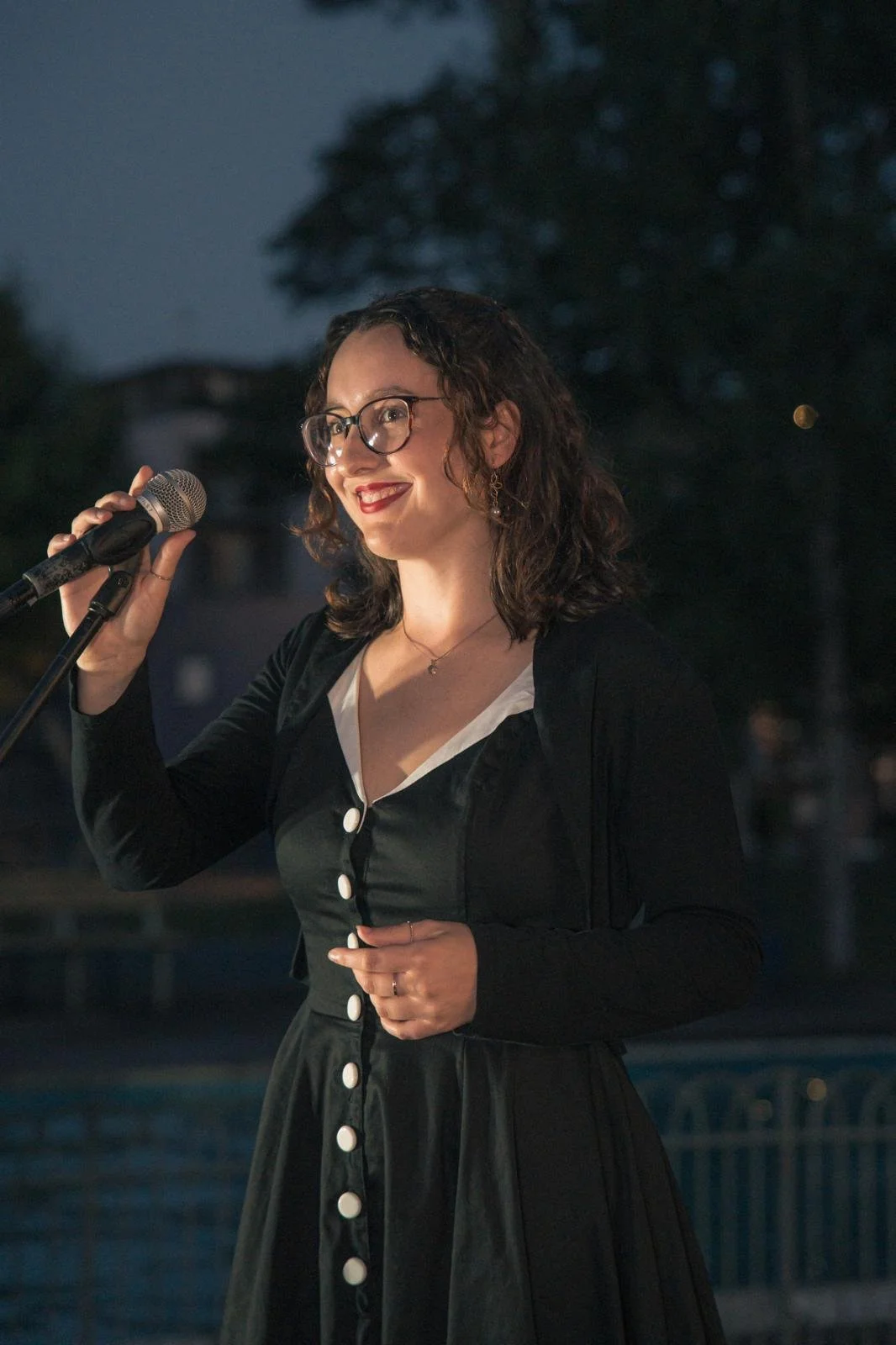 A young woman with curly brown hair and glasses smiling while holding a microphone on an outdoor stage during the evening.