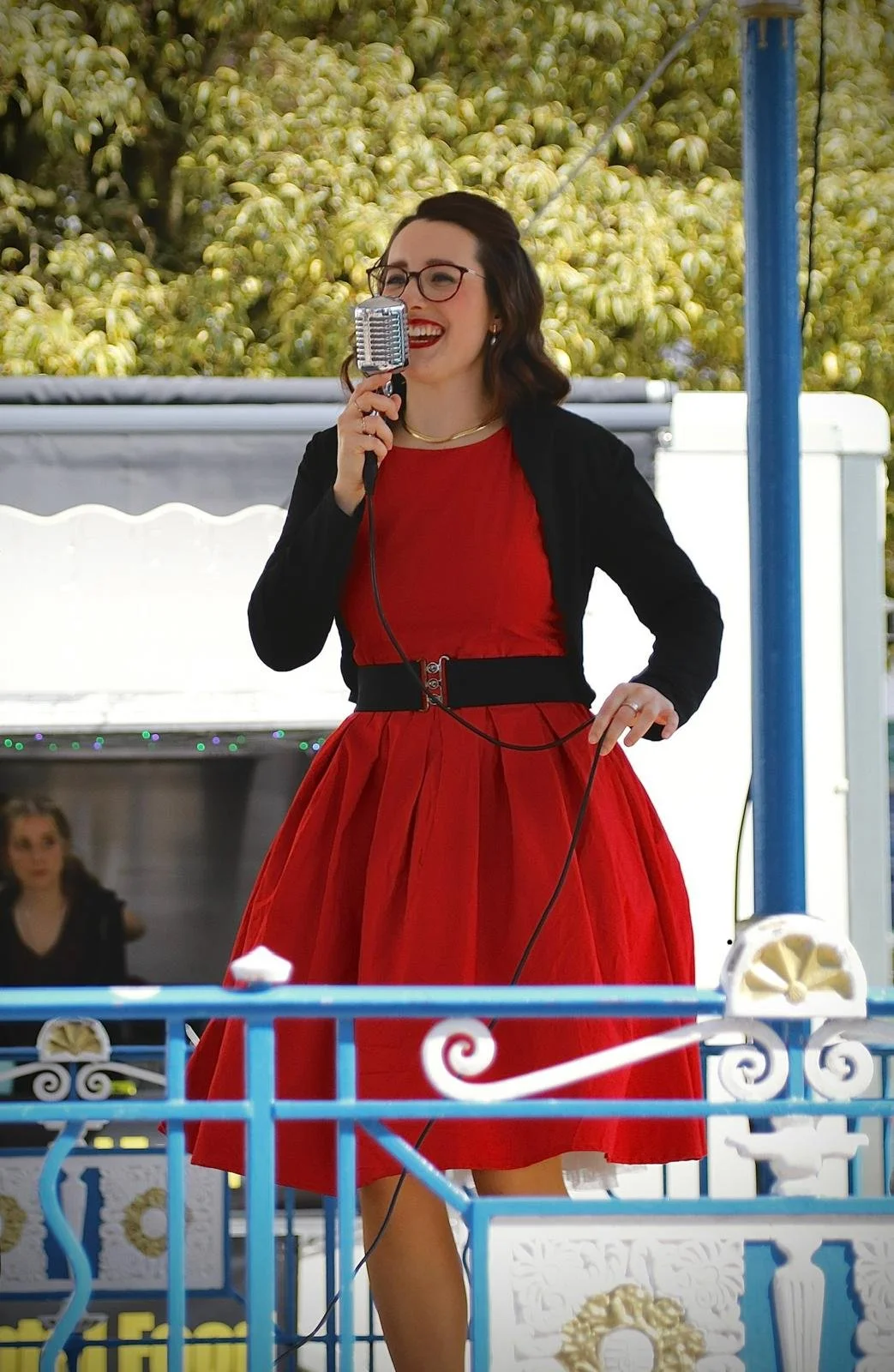 A woman wearing glasses, a red dress, and a black jacket singing into a vintage microphone outdoors, with a backdrop of green trees.