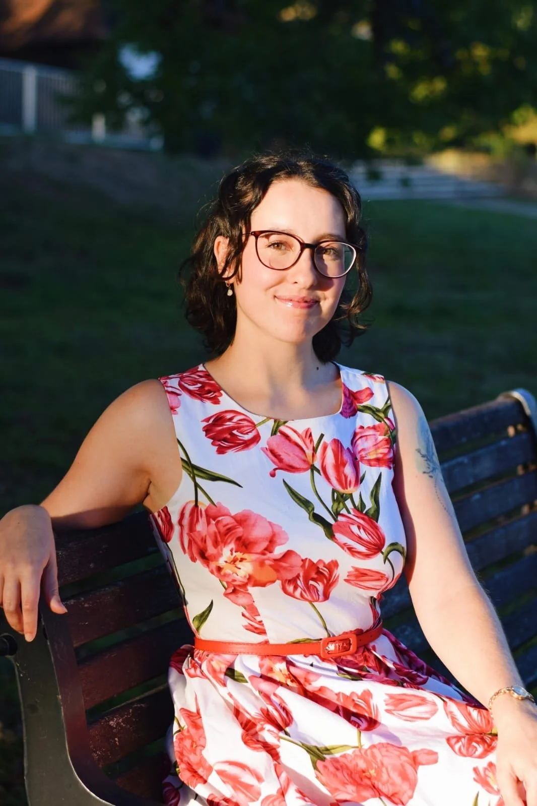 Keziah vintage jazz vocalist with glasses and short dark hair sitting on a park bench in sunlight, wearing a white dress with pink and red floral pattern and a red belt, smiling at the camera.