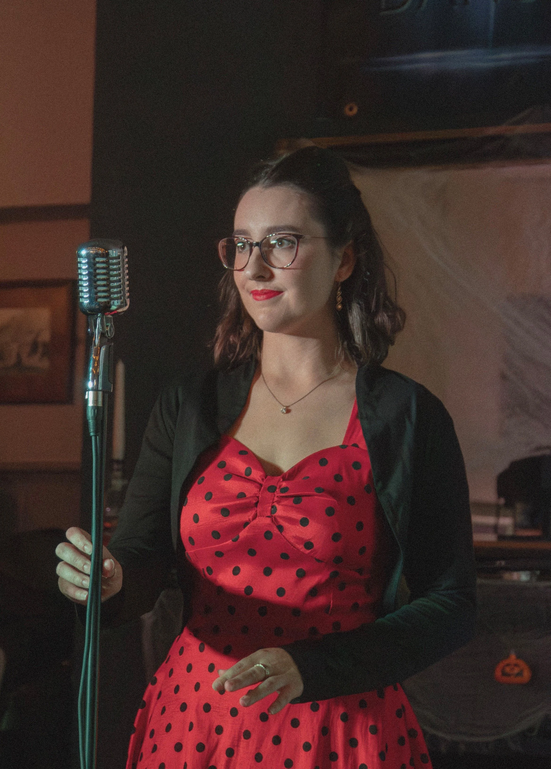 A woman with glasses, red lipstick, and earrings standing near a vintage microphone, wearing a red polka-dot dress with a black jacket in a dimly lit room.