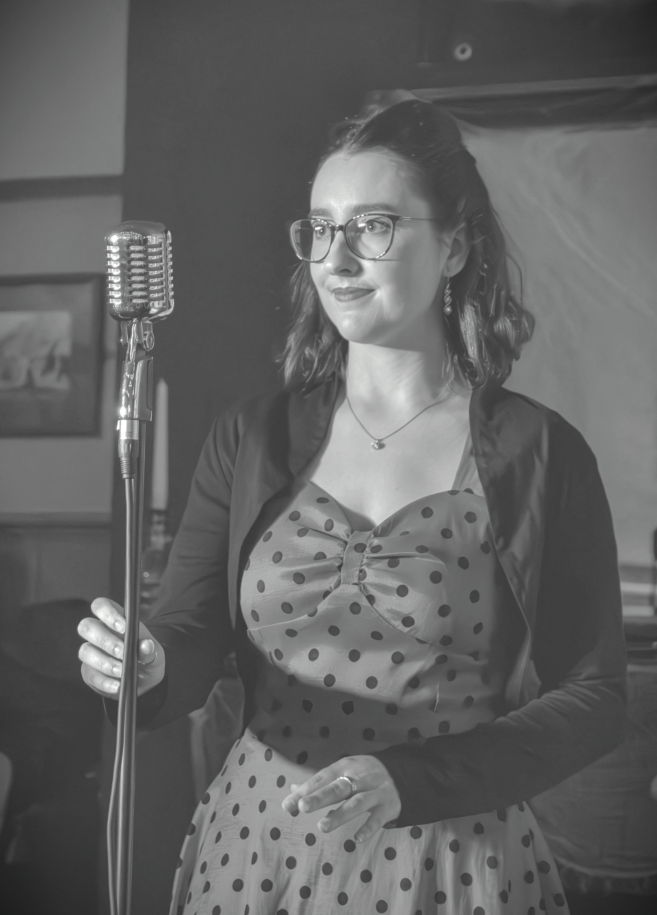 A woman in glasses and vintage style clothing stands near a microphone, holding the stand with her left hand. She has shoulder-length wavy hair and wears earrings and a necklace, smiling slightly.