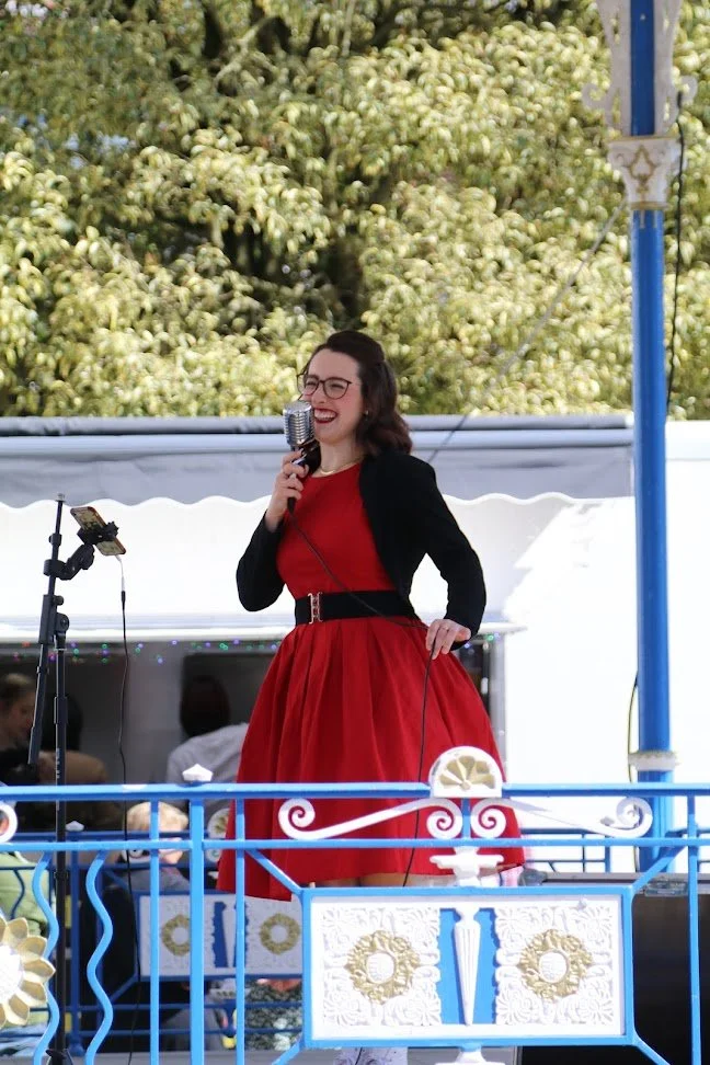Woman dressed in red and black, singing into a microphone on an outdoor stage with a decorative carousel railing, trees in the background, and people seated behind her.