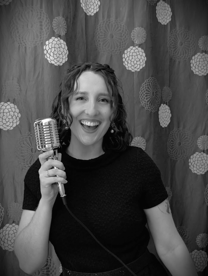 Woman with curly hair smiling, holding a vintage microphone, standing in front of a decorative backdrop with circular paper fans.