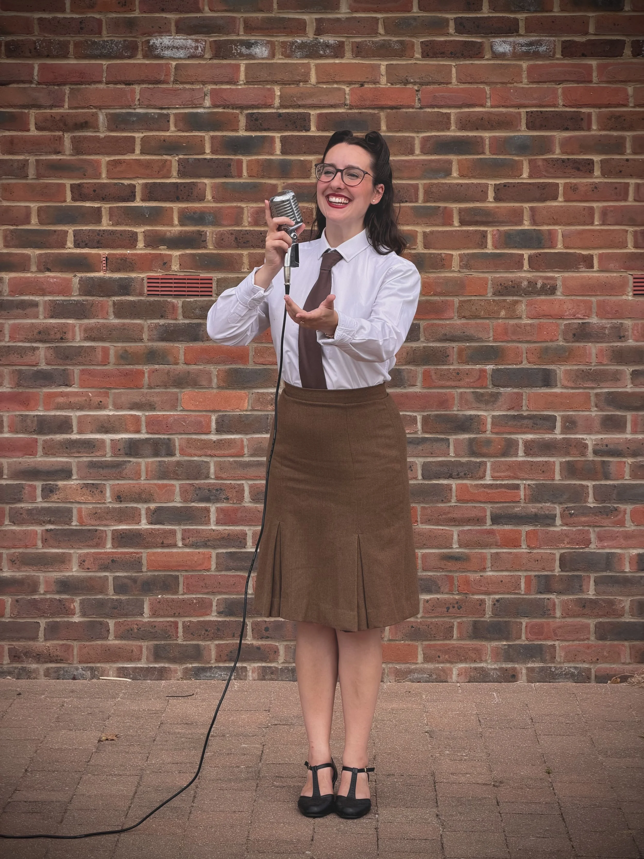 Keziah vintage jazz vocalist wearing glasses, a white shirt, a brown tie, and a brown skirt standing in front of a brick wall, holding a vintage microphone and smiling. She is wearing the ENSA (Entertainment Services Association) uniform that Dame Ve