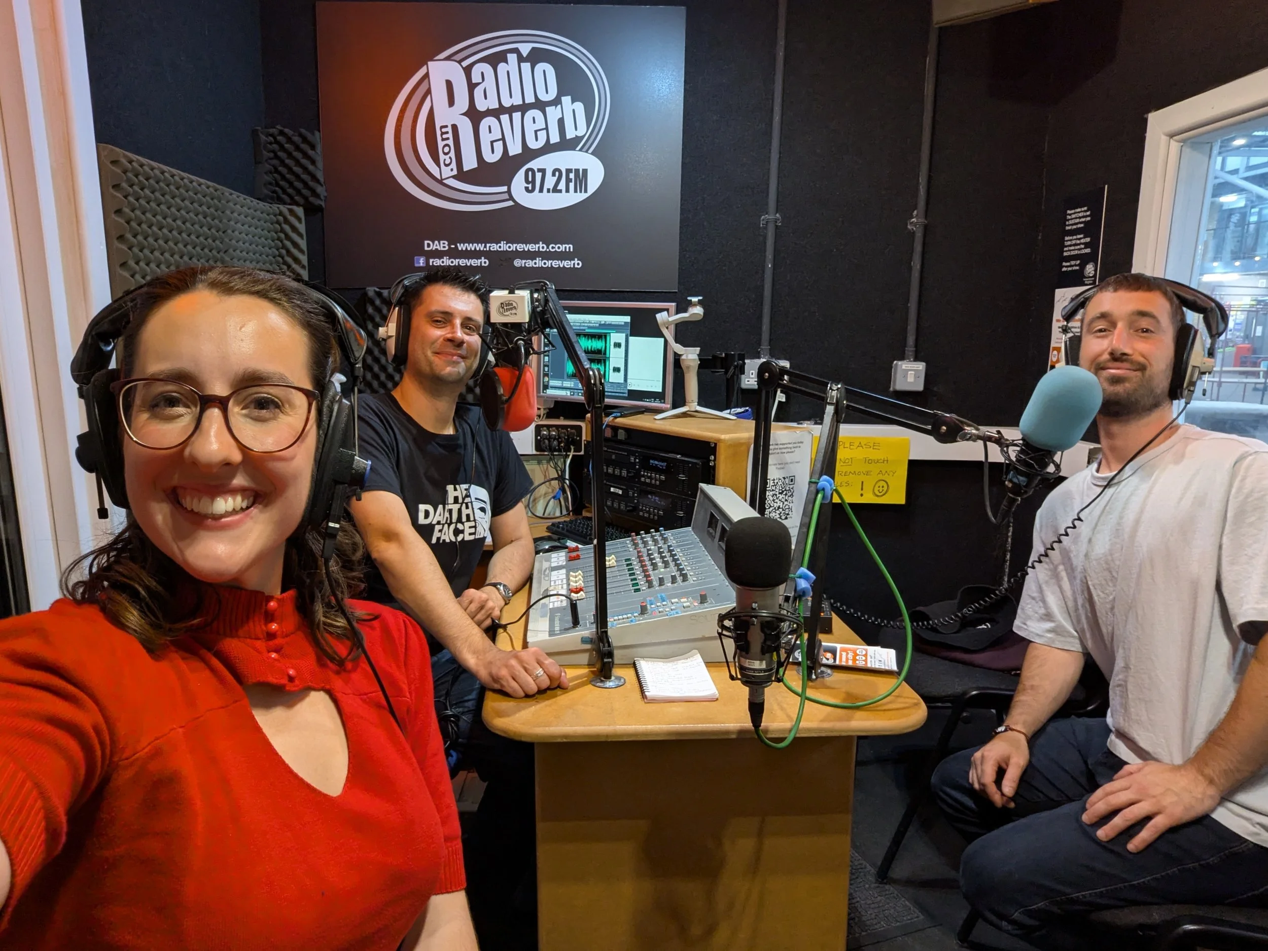 Four people are inside a radio studio with headphones and microphones. A woman in a red sweater is taking a selfie, with three men sitting behind her. The background features soundproofing panels and a logo on the wall reading 'Radio Reverb 97.2 FM', along with computer equipment and a yellow sign on the table.
