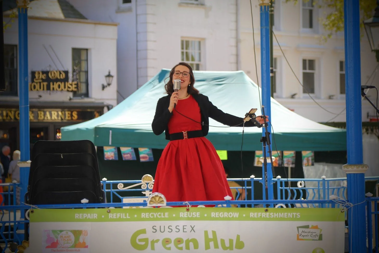 A woman in a red dress and black jacket speaking into a microphone on a stage at Sussex Green Hub during an outdoor event.