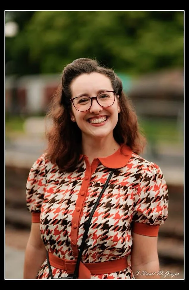 A smiling woman with glasses and wavy brown hair stands outdoors with a blurred background. She is wearing a patterned dress with shades of red, beige, black, and white, and has a camera strap around her neck.