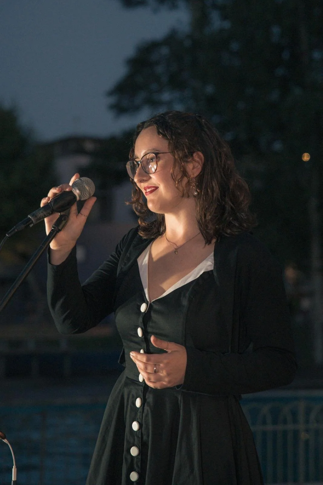 A woman with curly brown hair and glasses, wearing a black dress with white buttons and a white collar, is holding a microphone and speaking at an outdoor event in the evening.