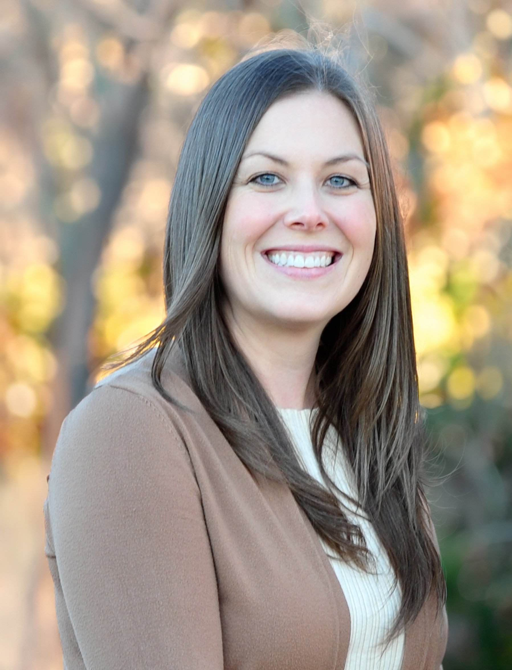 A young woman with long brown hair and blue eyes smiling outdoors with trees in the background