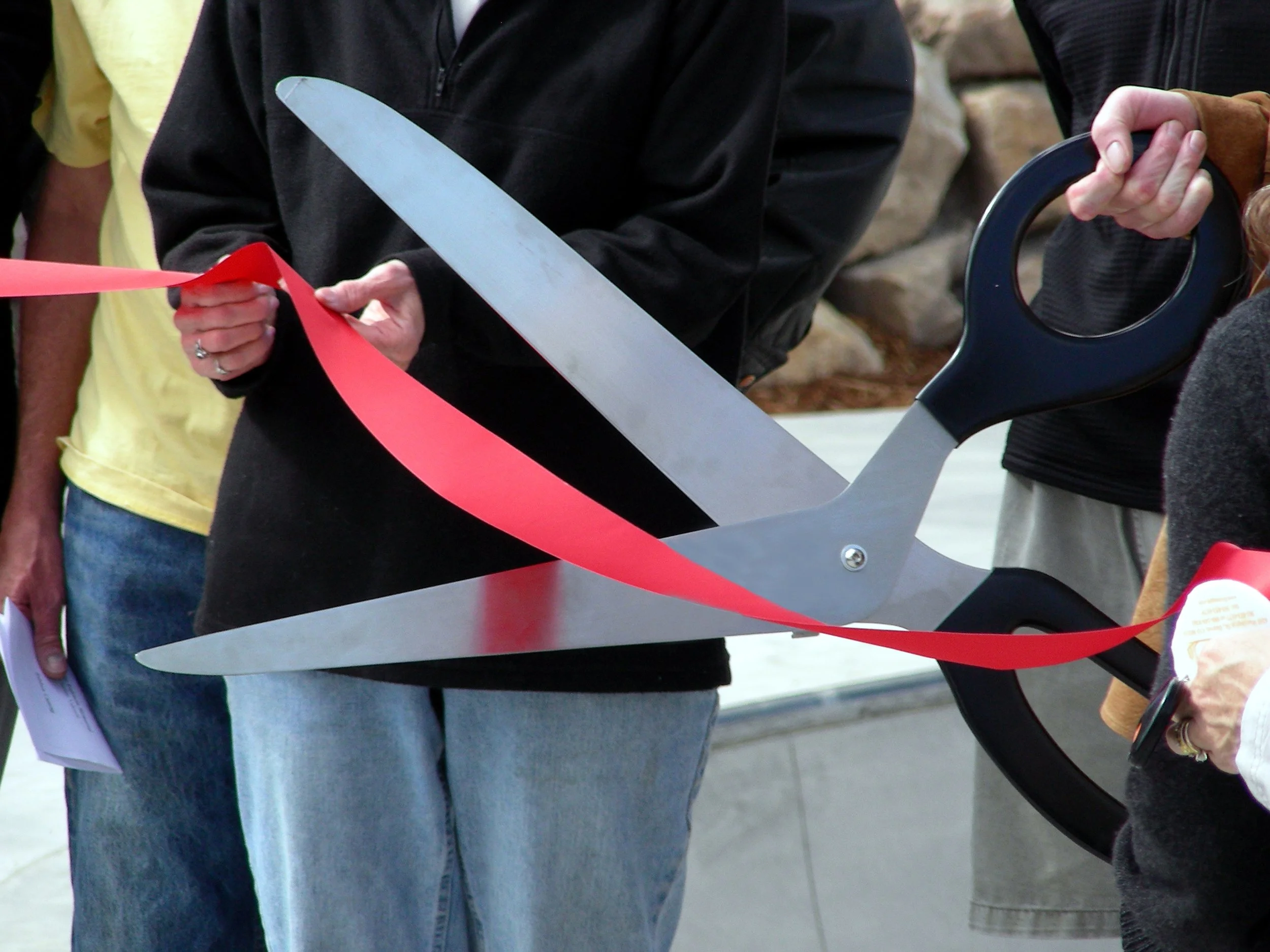 People at a ribbon-cutting event holding scissors and a tape, with some holding papers, outside with a stone wall in the background.