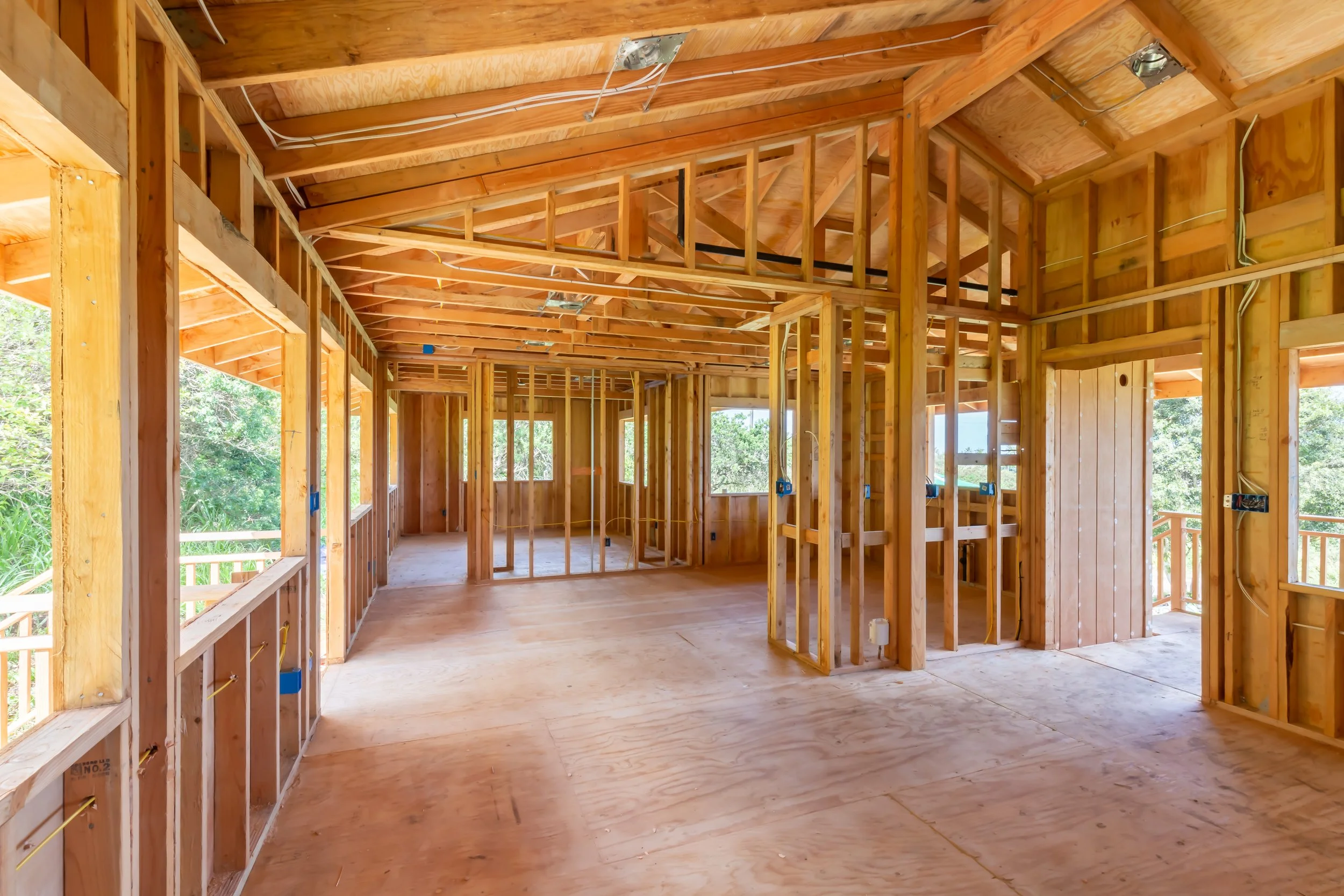 Interior view of a house under construction showing wooden framing and exposed ceiling with electrical wiring, with windows facing outdoors.