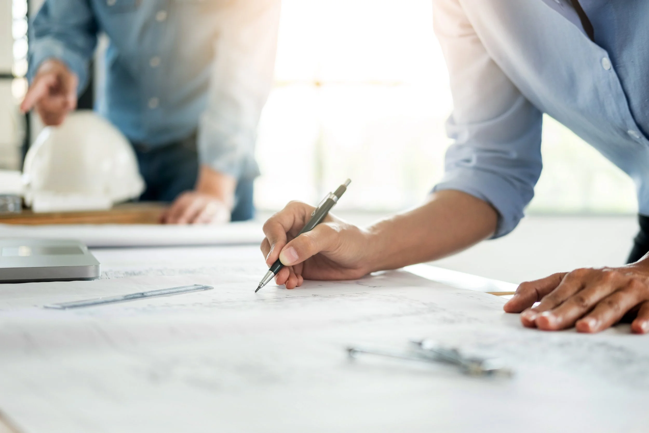 People working on blueprints or plans on a table with a person writing with a pen in the foreground.