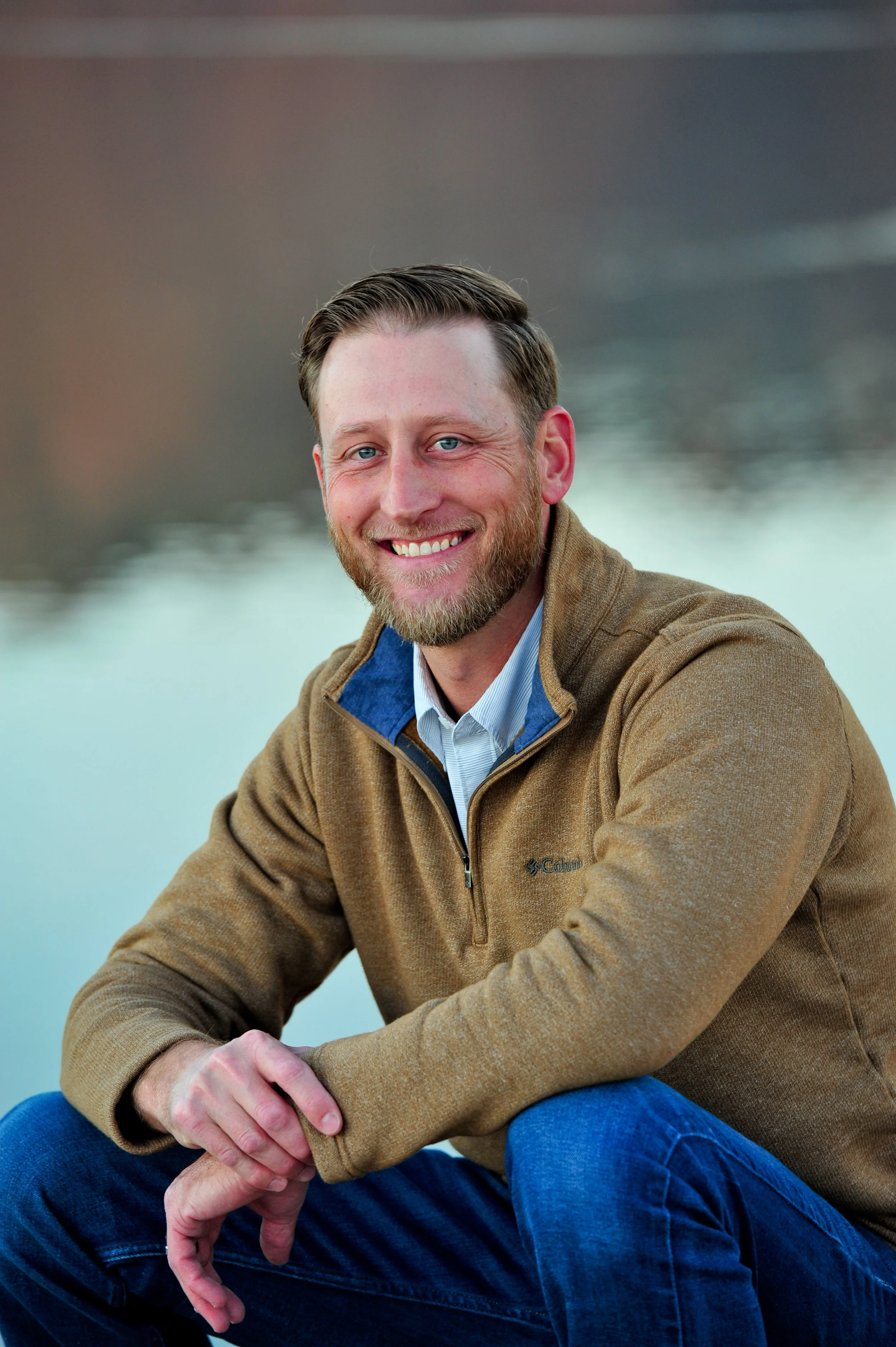 A smiling man with light skin, blue eyes, and short brown hair, wearing a tan Columbia fleece jacket and blue jeans, sitting outdoors by water with a blurred natural background.
