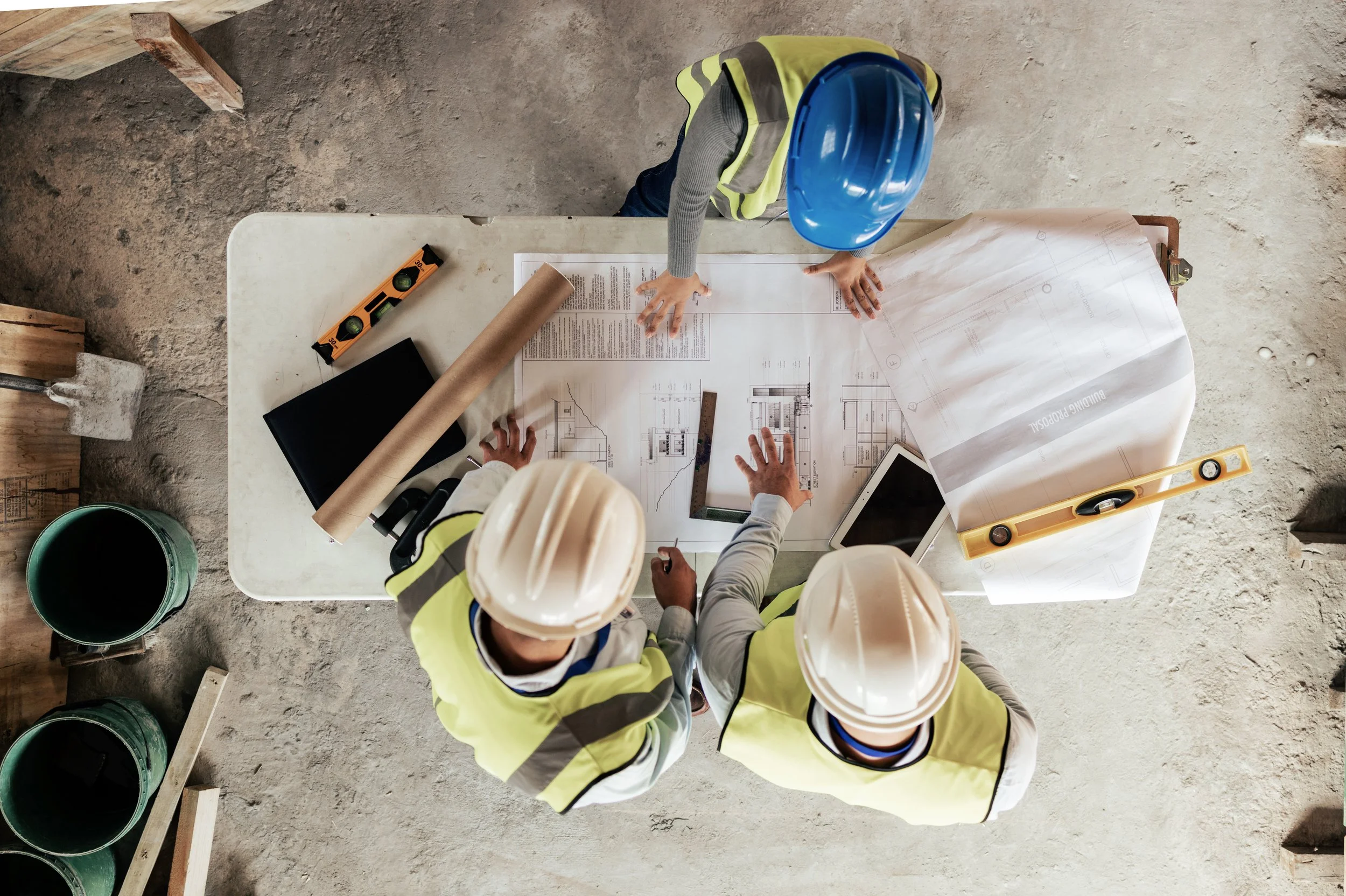 Two construction workers wearing yellow safety vests and white hard hats discussing blueprints on a construction site. Construction materials and tools are visible on the table and around them.