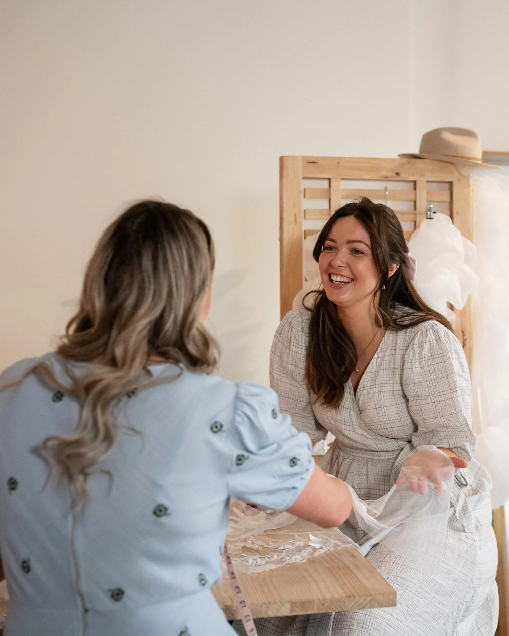 Two women smiling and talking in a room with a wooden bed frame, one of them is seated, and the other week leaning forward with her arm extended.