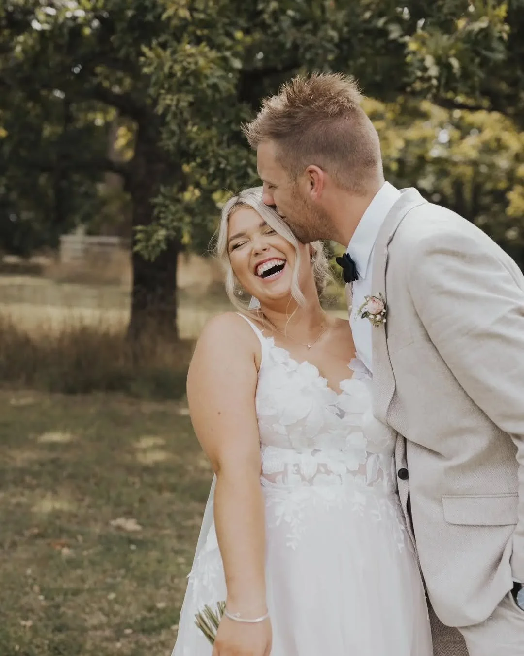 A joyful bride with blonde hair in a white wedding dress laughing as a groom in a light gray suit kisses her forehead outdoors with trees in the background.
